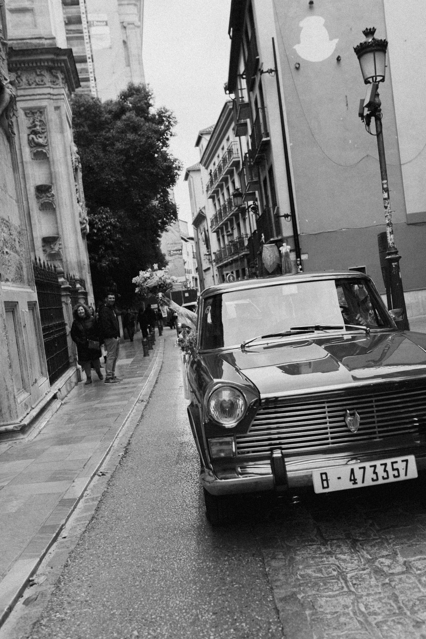 Une rue européenne avec des bâtiments résidentiels et commerciaux, un lampadaire, une voiture ancienne garée sur le trottoir, et des personnes marchant, dont une tenant un bouquet de fleurs.