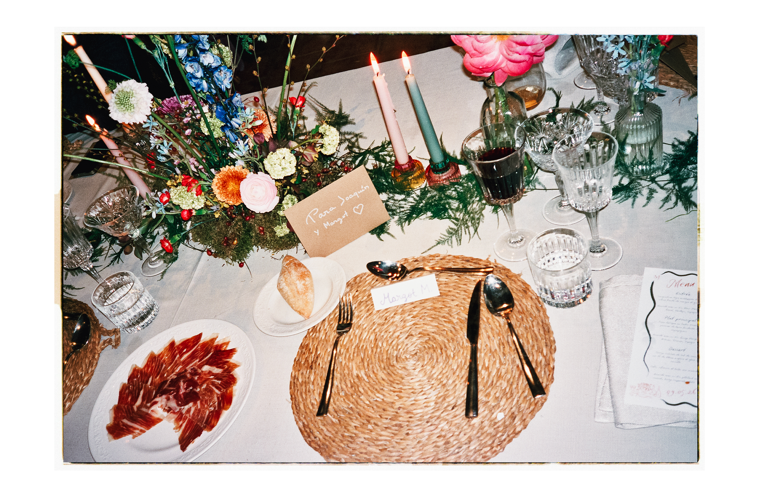 Table de dîner décorée avec un centre de fleurs, bougies allumées, verres de vin rouge, assiettes de charcuterie, et une note écrite à la main. Décorations florales colorées avec des bougies roses et vertes, assiettes en verre fin, et une nappe blanc