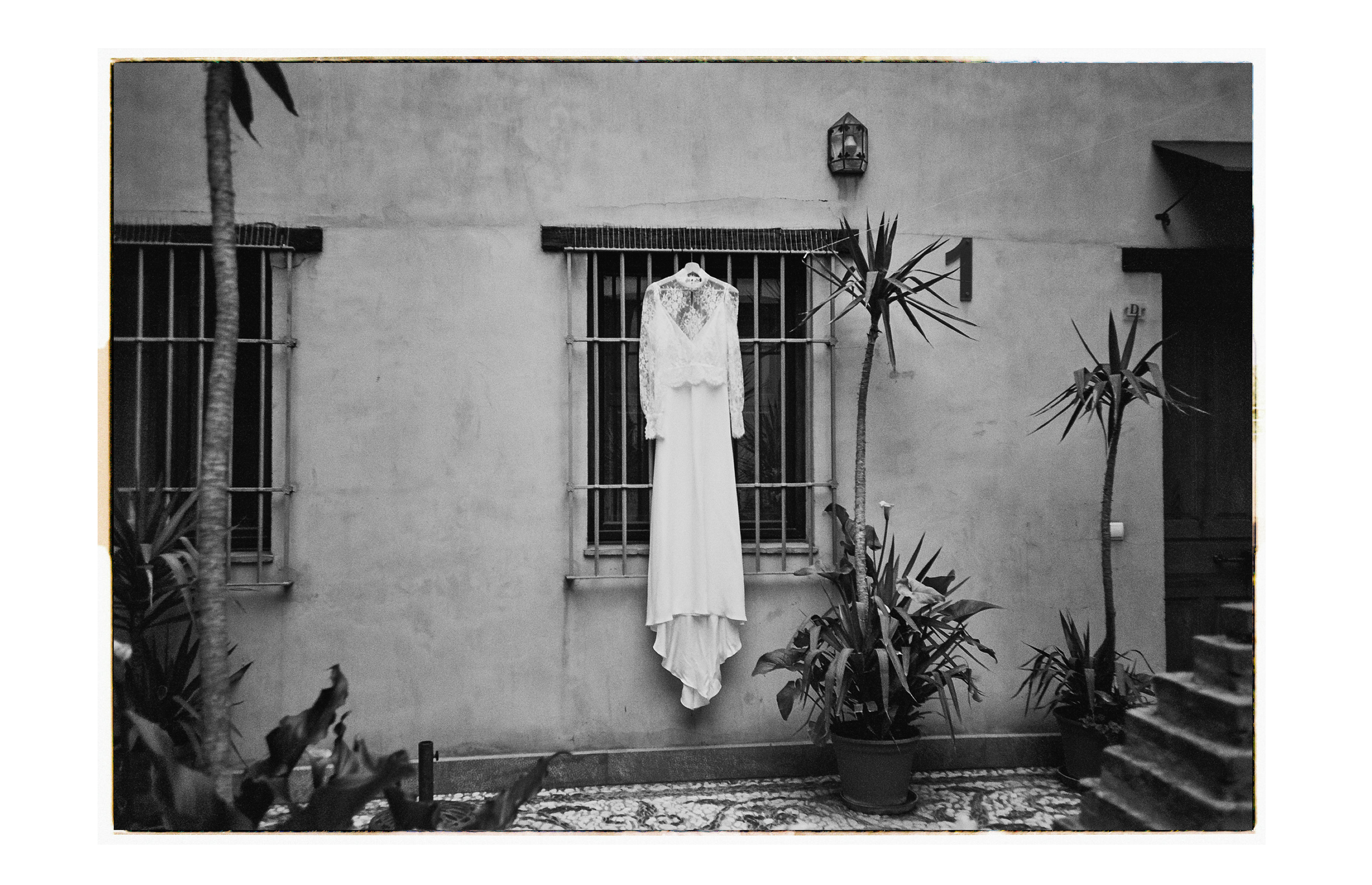 Une robe de mariée blanche suspendue à une fenêtre avec barreaux, entourée de plantes en pot dans une cour intérieure, en noir et blanc.