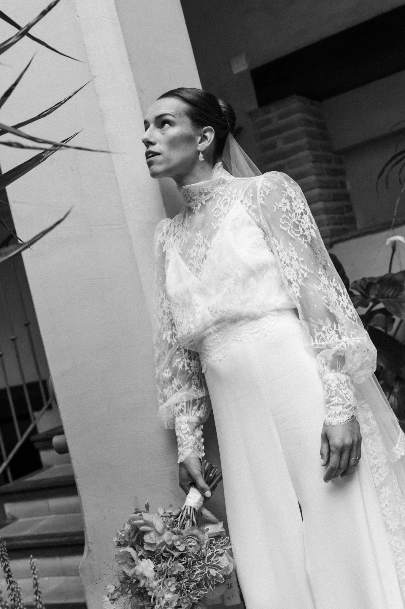 Une femme en robe de mariage en dentelle, portant un voile, tenant un bouquet de fleurs, photographiée en noir et blanc à l'intérieur, près d'un mur avec des briques visibles.