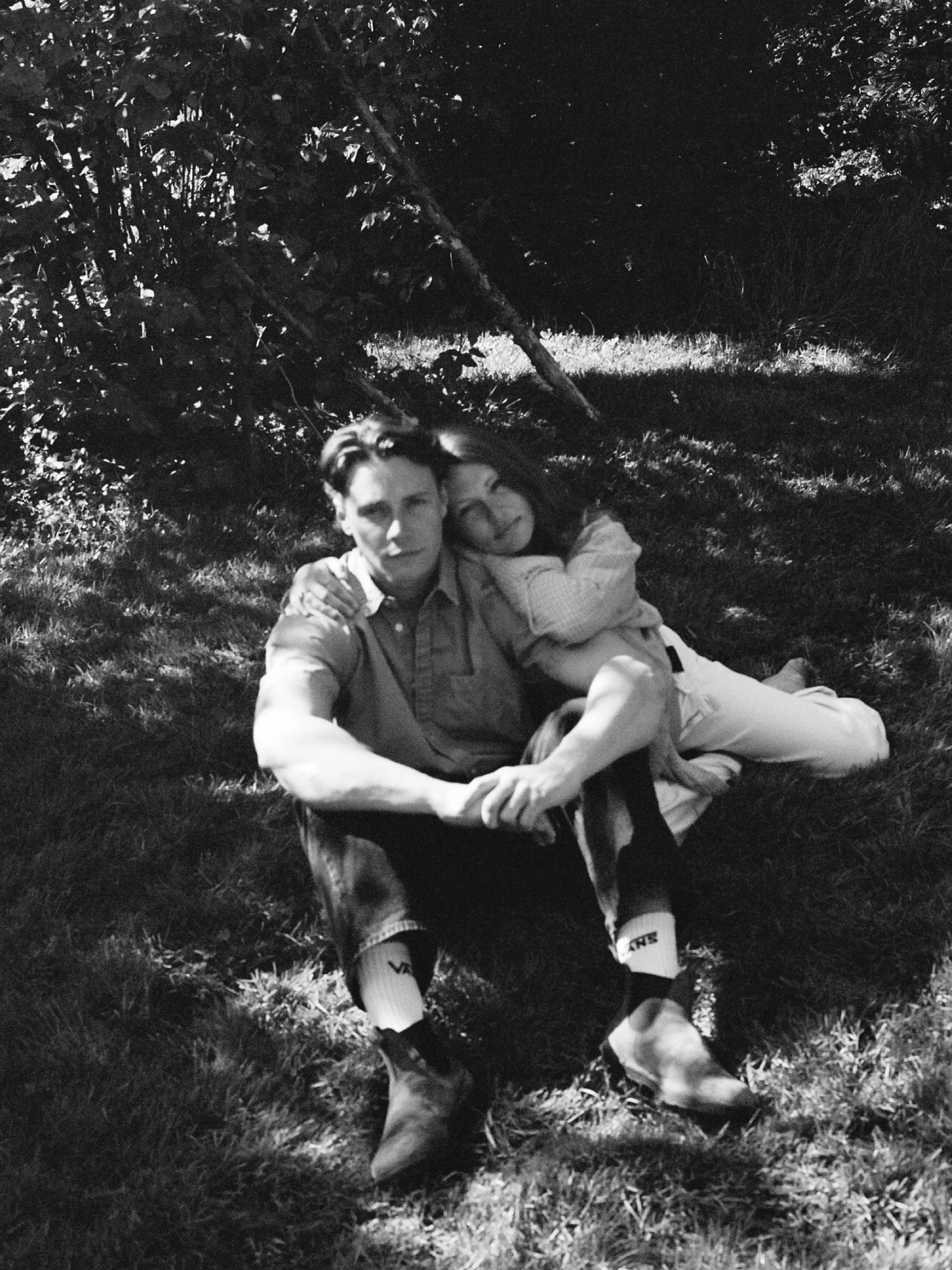 Un homme et une femme assis sur l'herbe dans un jardin la nuit, prenant un selfie. L'homme regarde l'objectif avec une expression neutre, la femme sourit, elle repose sa tête sur l'épaule de l'homme. La scène est en noir et blanc.