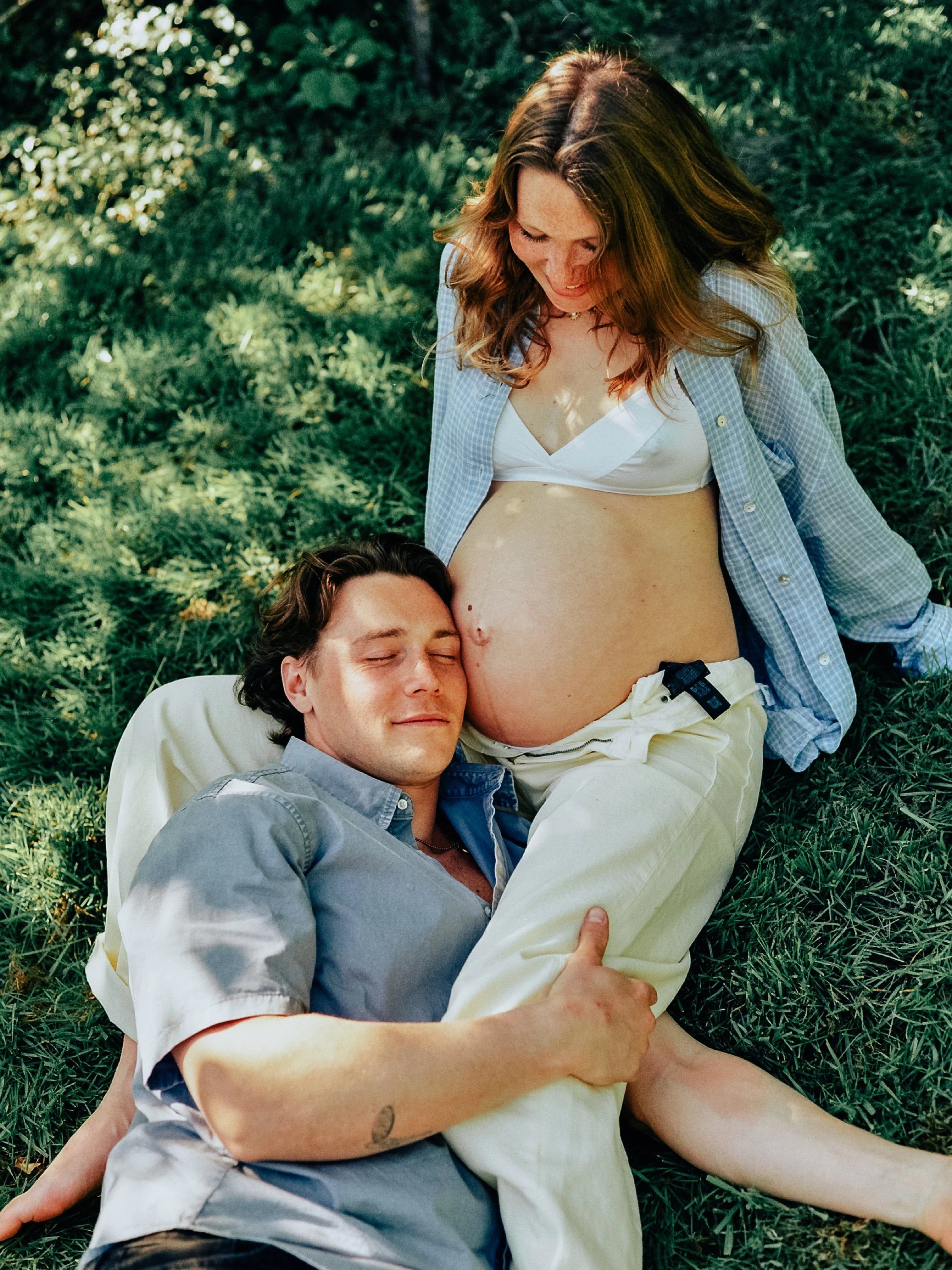 Un couple, un homme et une femme, allongés sur l'herbe dans un parc. La femme est enceinte, elle sourit et regarde son ventre, tandis que l'homme repose la tête sur sa jambe, tous deux semblent heureux et complices.