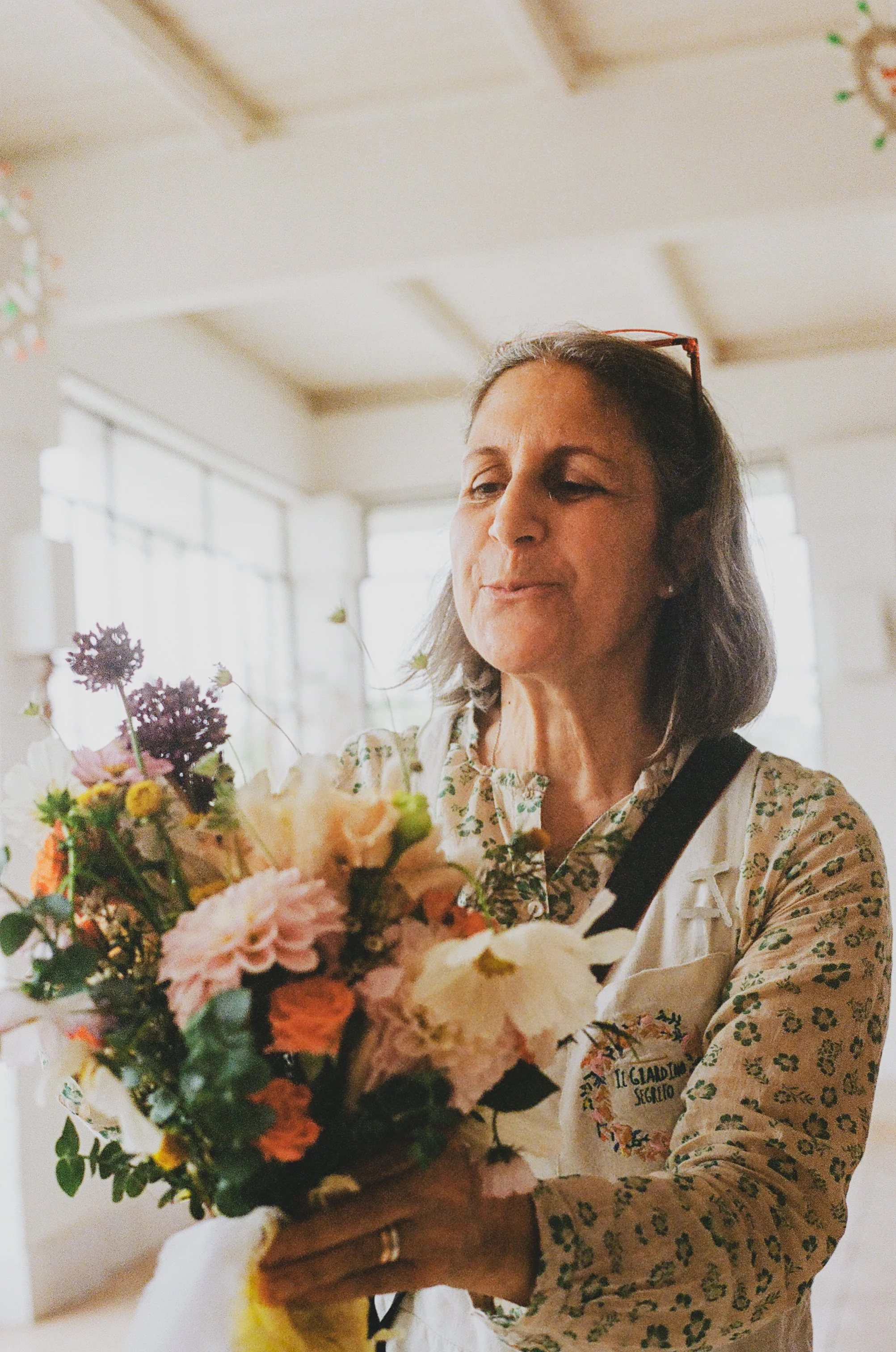 Une femme blonde portant une chemise à fleurs et portant des lunettes rouges, tenant un bouquet de fleurs colorées dans un intérieur lumineux.