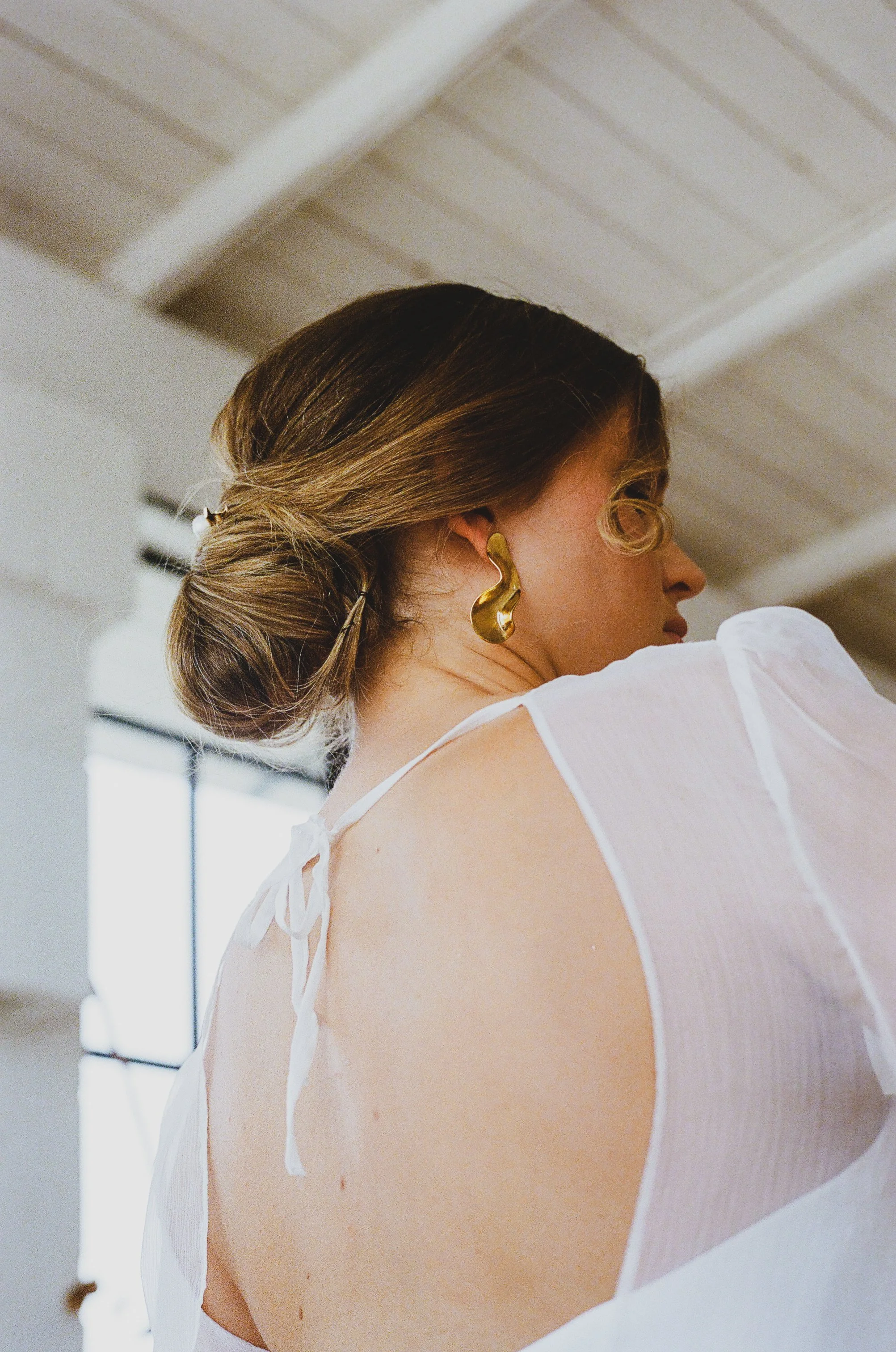 Une femme vue de profil avec une coiffure attachée en chignon et portant des boucles d'oreilles en forme de vague, portant une blouse blanche avec des bretelles. Le décor intérieur est lumineux avec un plafond en bois et des fenêtres en arrière-plan.