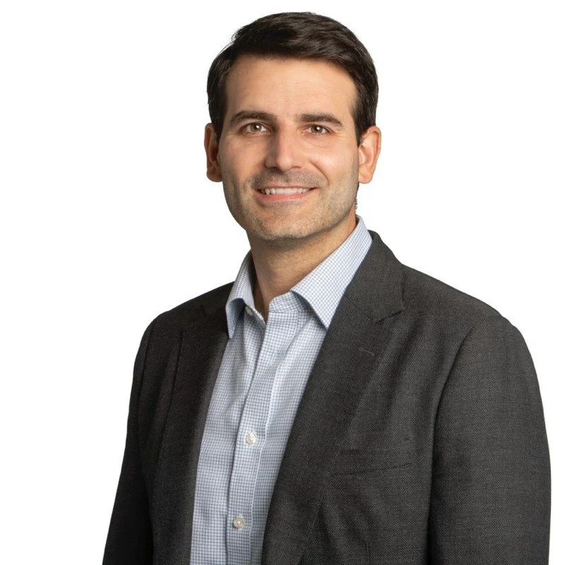 A professional man in a dark gray suit and light blue collared shirt smiling in front of a white background.