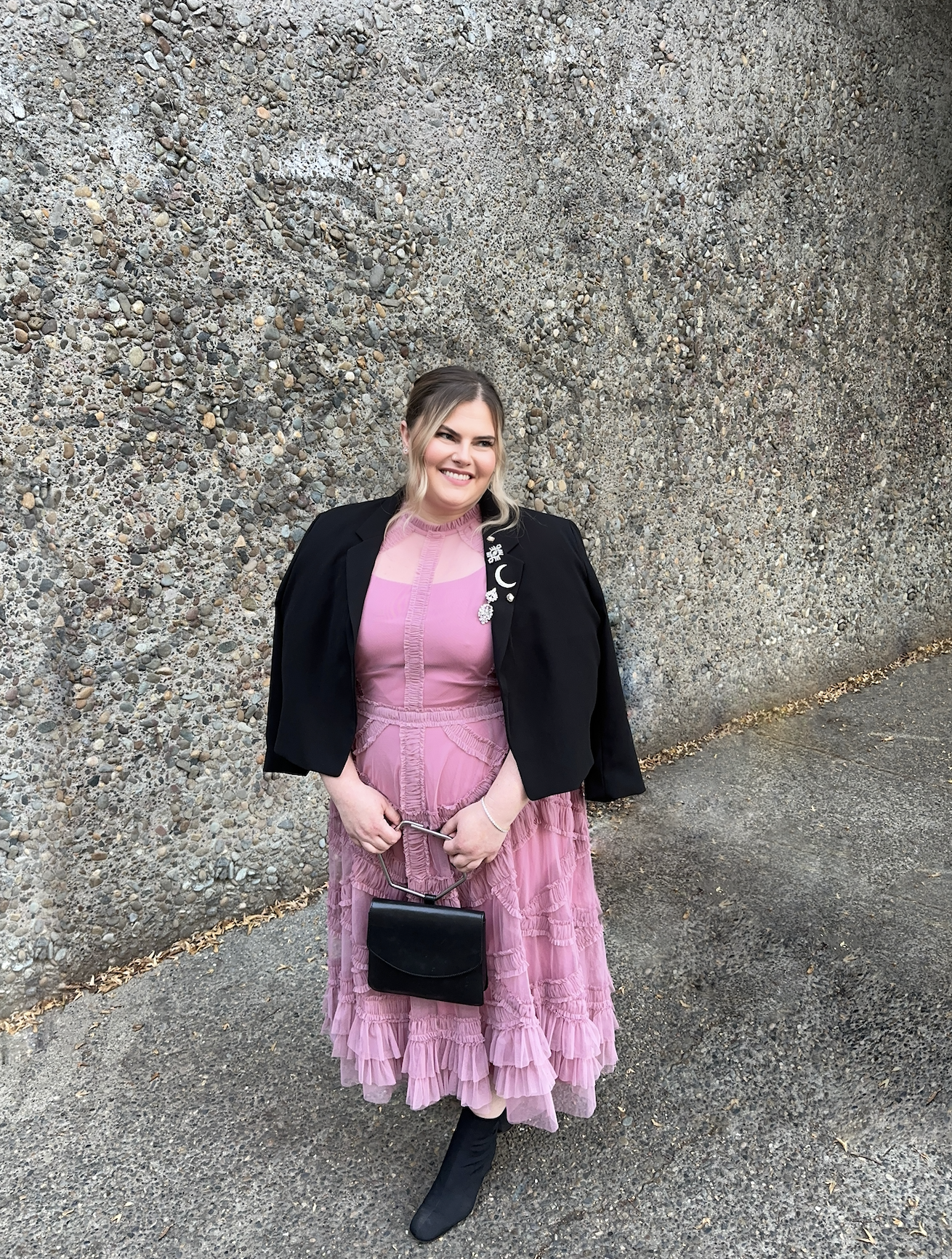 A woman in a pink ruffled dress and black boots standing in front of a textured stone wall, holding a small black handbag.