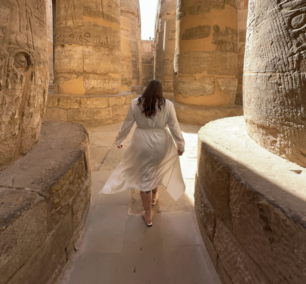 A woman in a white dress walking through ancient stone columns with carved hieroglyphics, resembling an archaeological site, possibly an Egyptian temple.