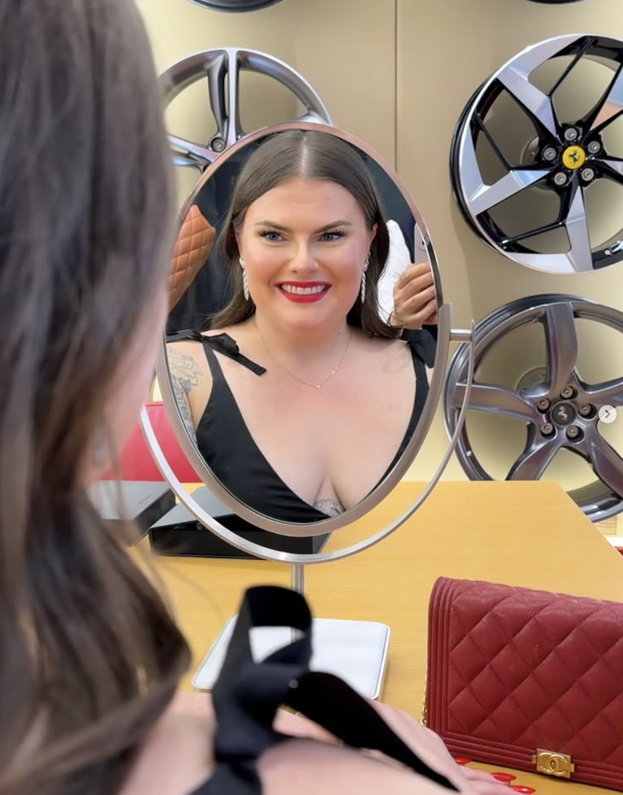 A woman with dark hair and red lipstick looking at her reflection in a small round mirror, surrounded by car rims hanging on a beige wall.