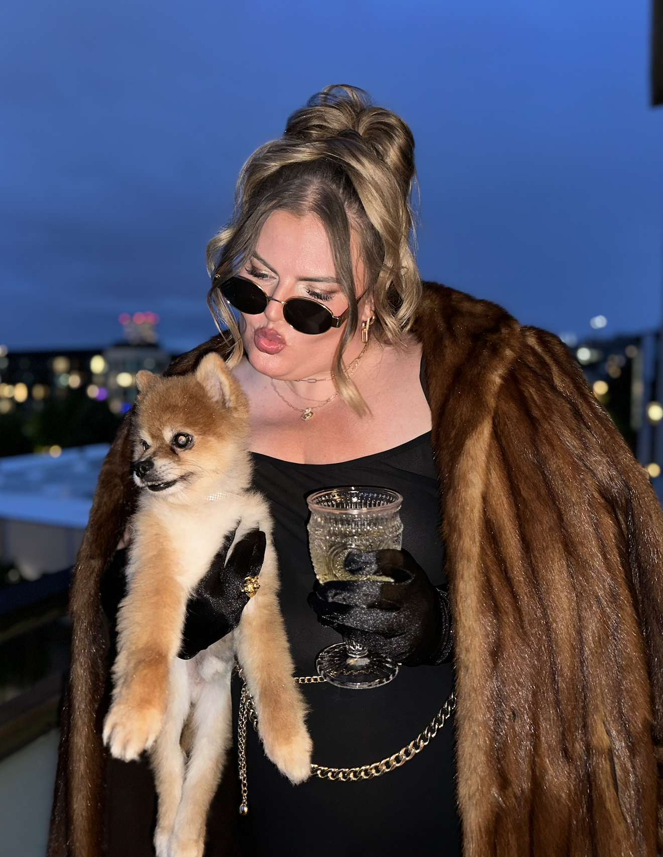 Woman with blonde hair wearing sunglasses, a fur coat, black gloves, and jewelry, holding a small puppy with a light brown and white coat, on a rooftop at night with city lights in the background.