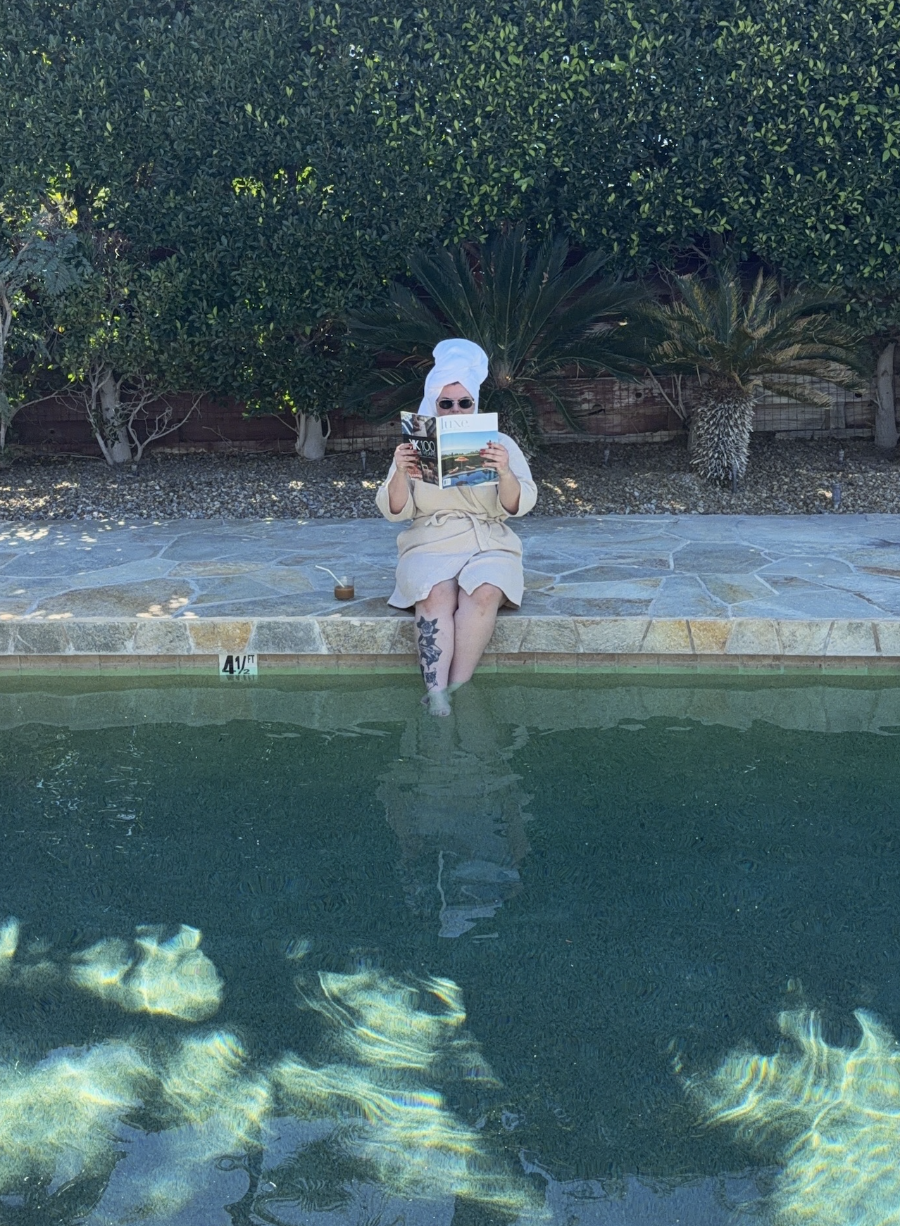 A woman sitting at the edge of a swimming pool reading a magazine. She is wearing sunglasses, a beige robe, and has a towel wrapped around her head. Her feet are submerged in the water, and there's a tattoo on her right leg. The background features tall green bushes and palm trees.