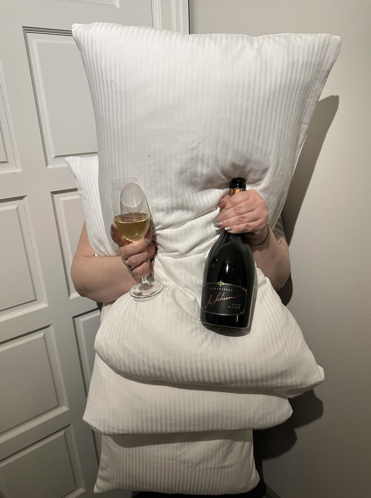 Person holding a large pillow in front of their face, holding a wine glass of white wine in one hand and a bottle of wine in the other, standing indoors near a white door.