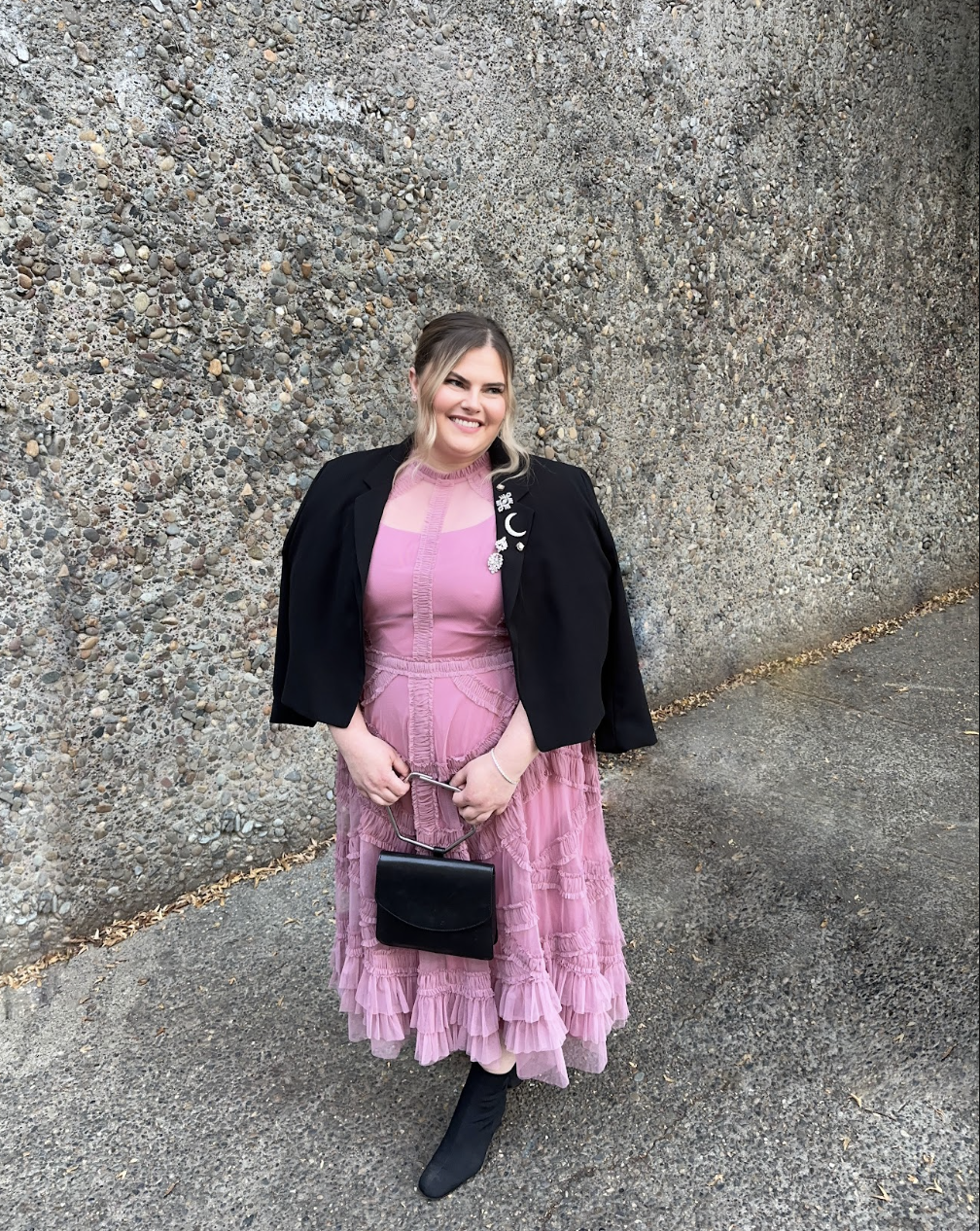 A woman standing outdoors in front of a textured concrete wall, smiling, wearing a pink layered dress, black boots, a black blazer with decorative pins, and holding a small black handbag.