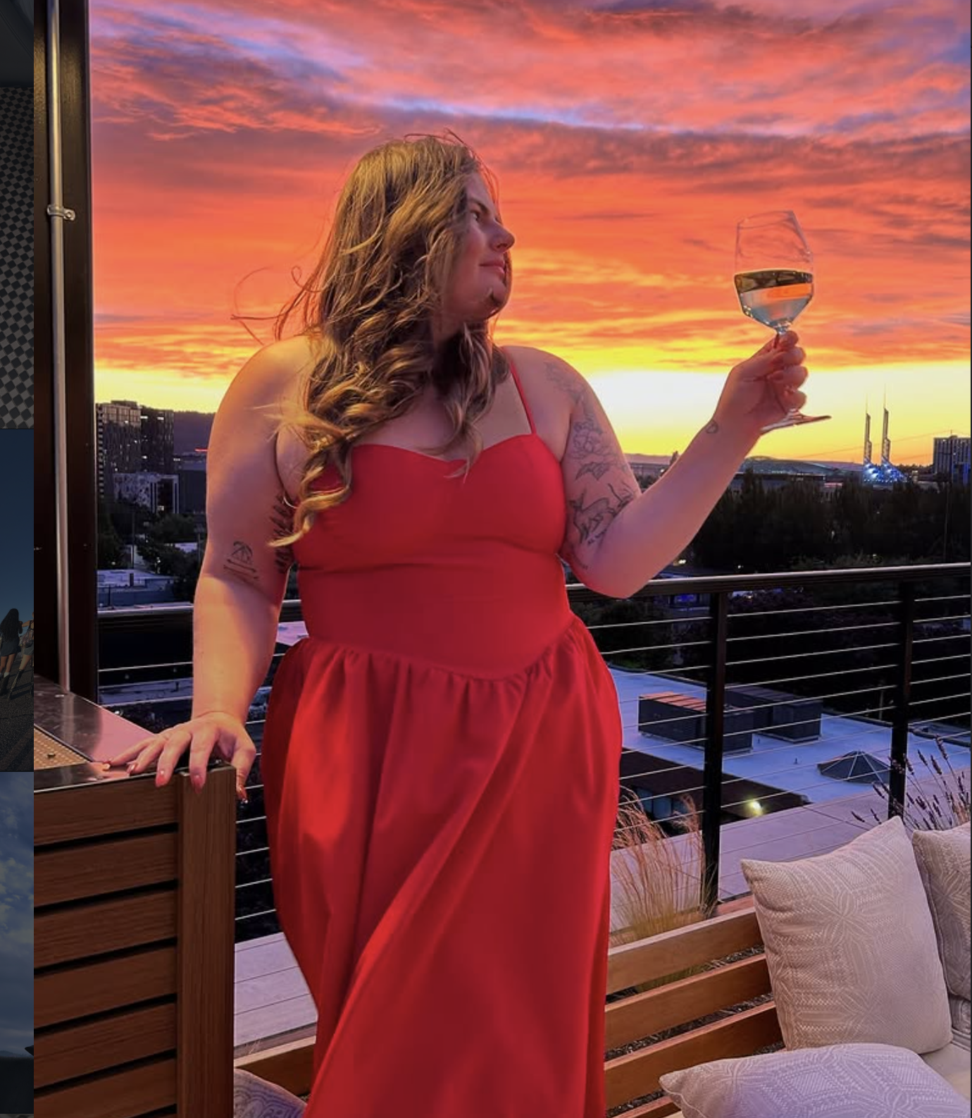 A woman in a red dress stands on a balcony during sunset, holding a glass of white wine and looking at the sky with colorful clouds and cityscape in the background.