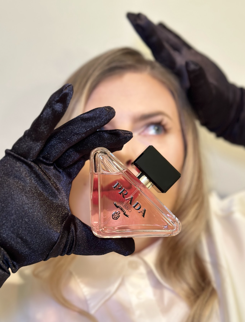 A woman with blond hair in black gloves holds a Prada Milano perfume bottle in front of her face.