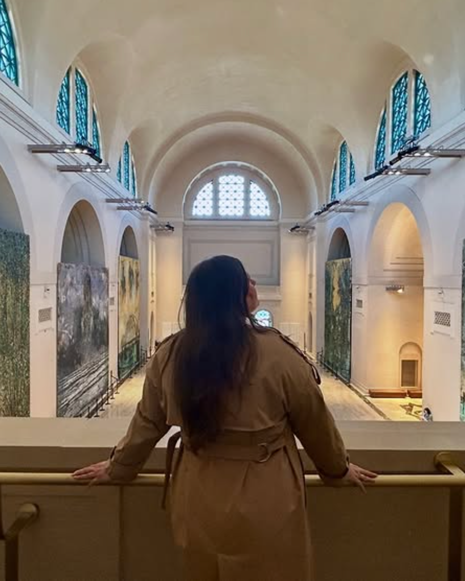 A woman in a tan trench coat with long dark hair is standing on a balcony in an art museum, looking at paintings displayed on the walls inside a large, bright gallery with high vaulted ceilings and blue window accents.