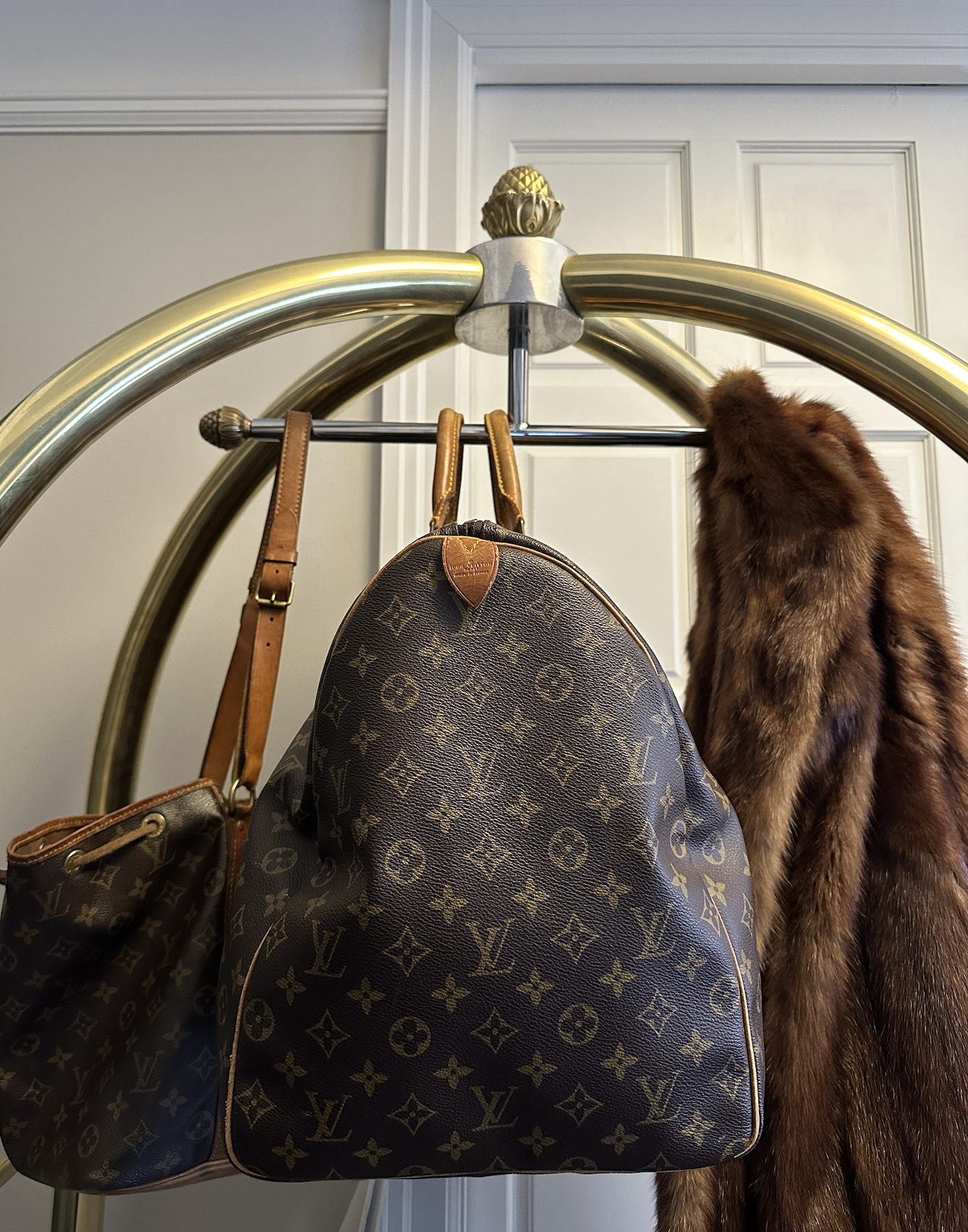 A luggage cart with three Louis Vuitton bags hanging on it, and a fur coat draped over the side of the cart, with a plain white wall and door in the background.