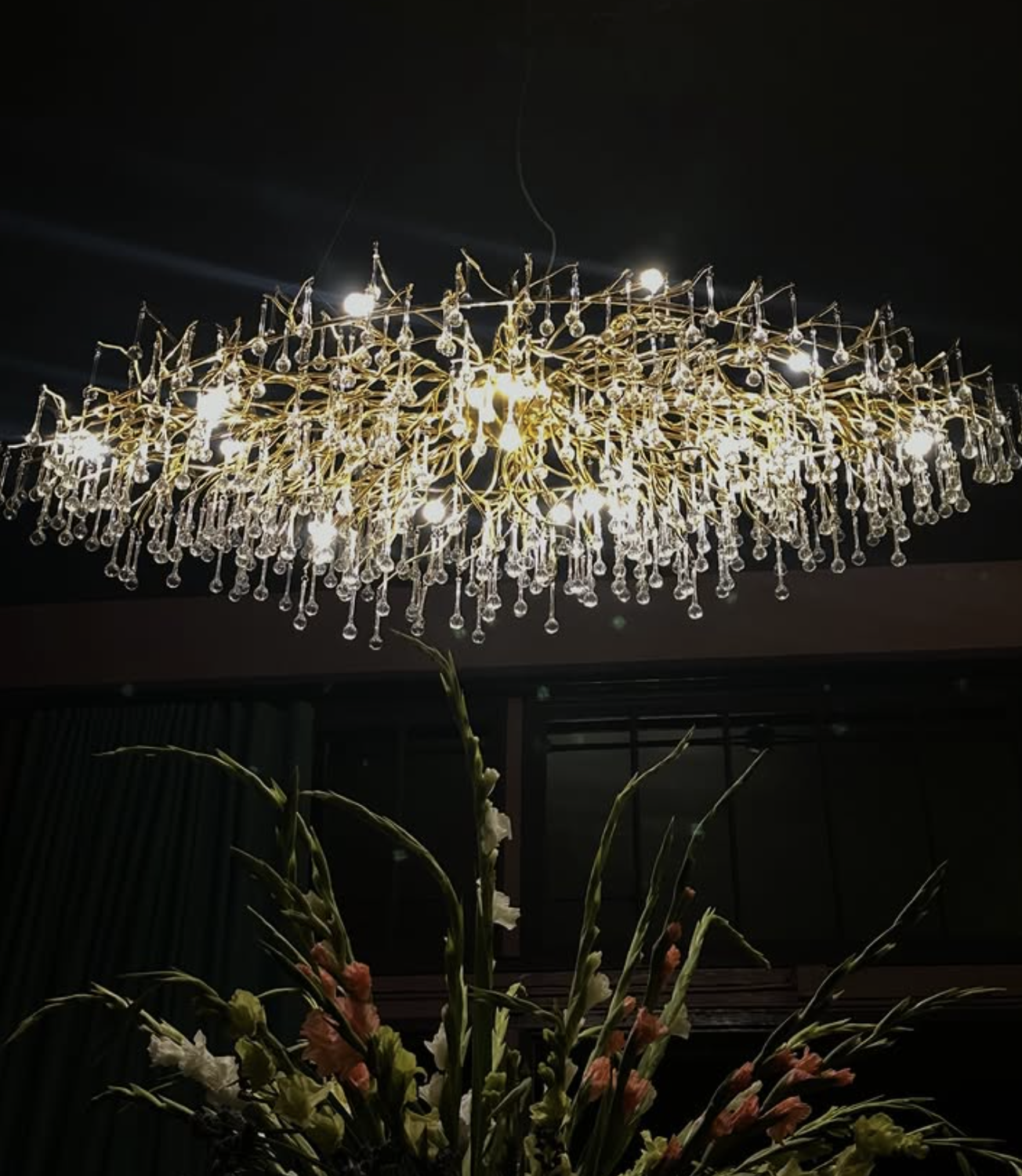 A luxurious chandelier with numerous crystal ornaments hangs from a dark ceiling above a vase with tall flowers.