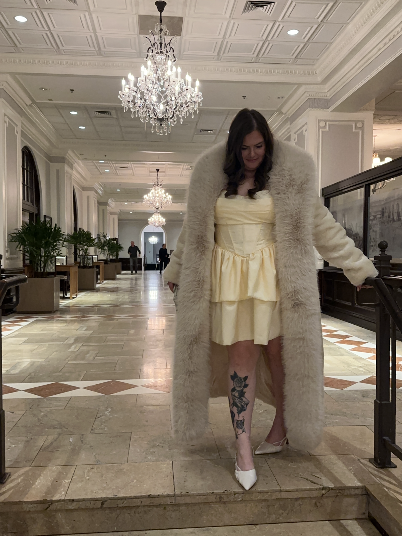 A woman in a beige dress with a large cream-colored fur coat standing in an elegant, well-lit hotel lobby, with chandeliers hanging from the ceiling, plants along the walls, and marble flooring.