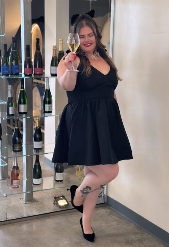 A woman dressed in a black dress and black heels, smiling and holding a glass of champagne, standing next to a display of wine bottles in a modern setting.