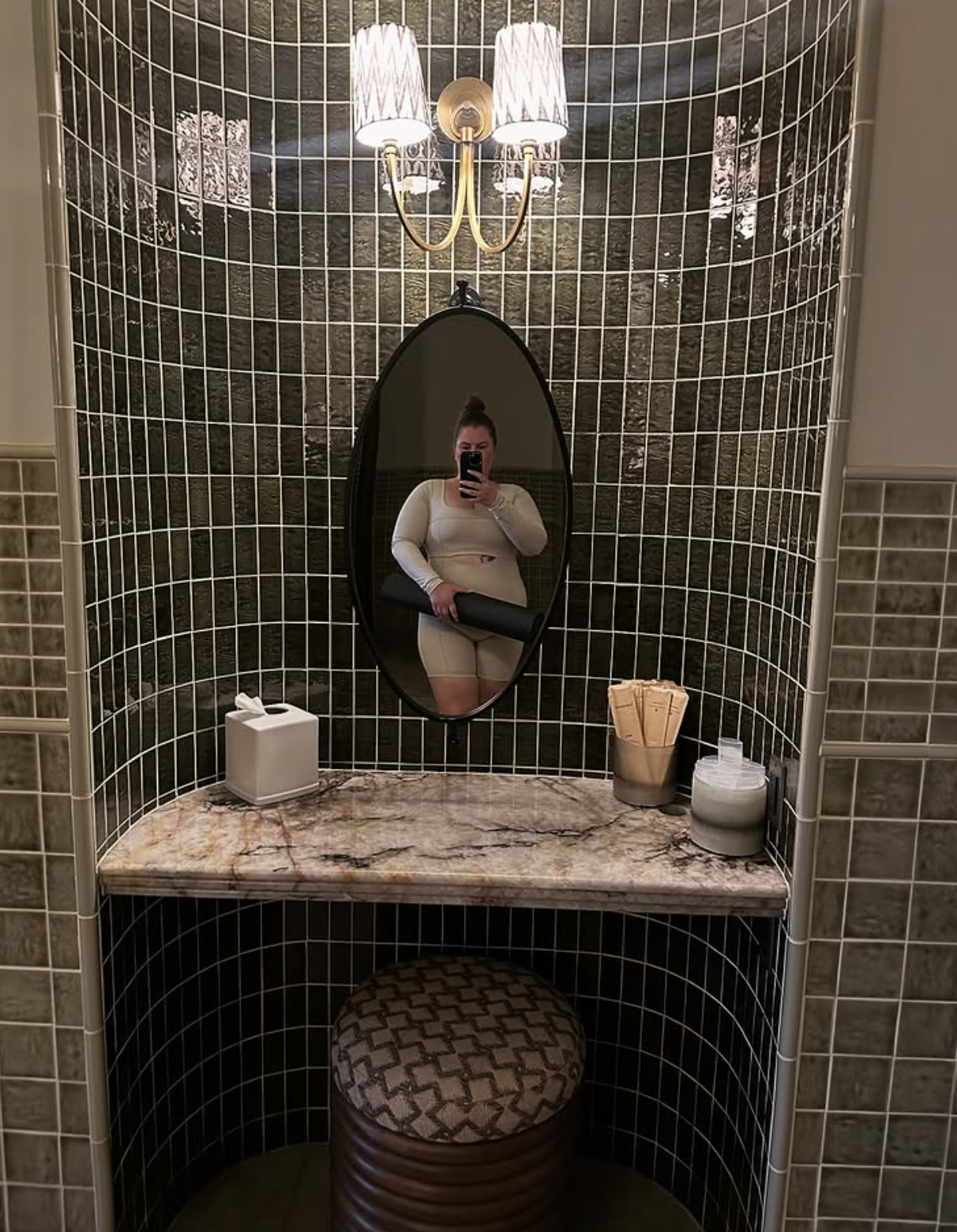 A bathroom with black tiled walls, a marble countertop, and a vertical oval mirror. A person taking a selfie is visible in the mirror, holding a black rolled mat, wearing a cream-colored long sleeve top and fitted shorts. On the countertop, there is a tissue box, a cup holding paper towels, and a container with toiletries. A stool with a patterned cushion is underneath the countertop, and a wall-mounted light fixture with two lamps is above the mirror.
