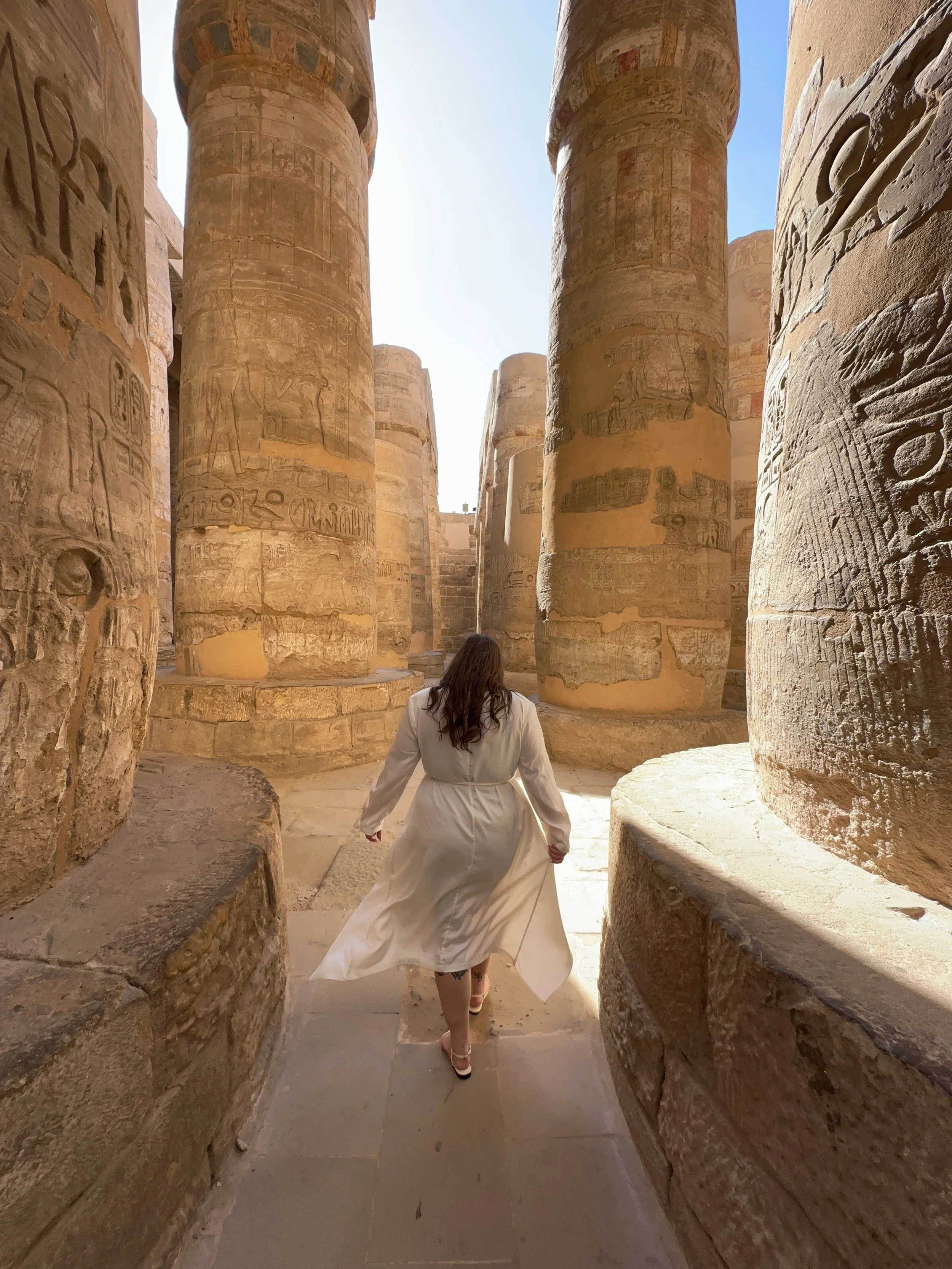 A woman in a white coat walking through ancient Egyptian temple columns decorated with hieroglyphics.