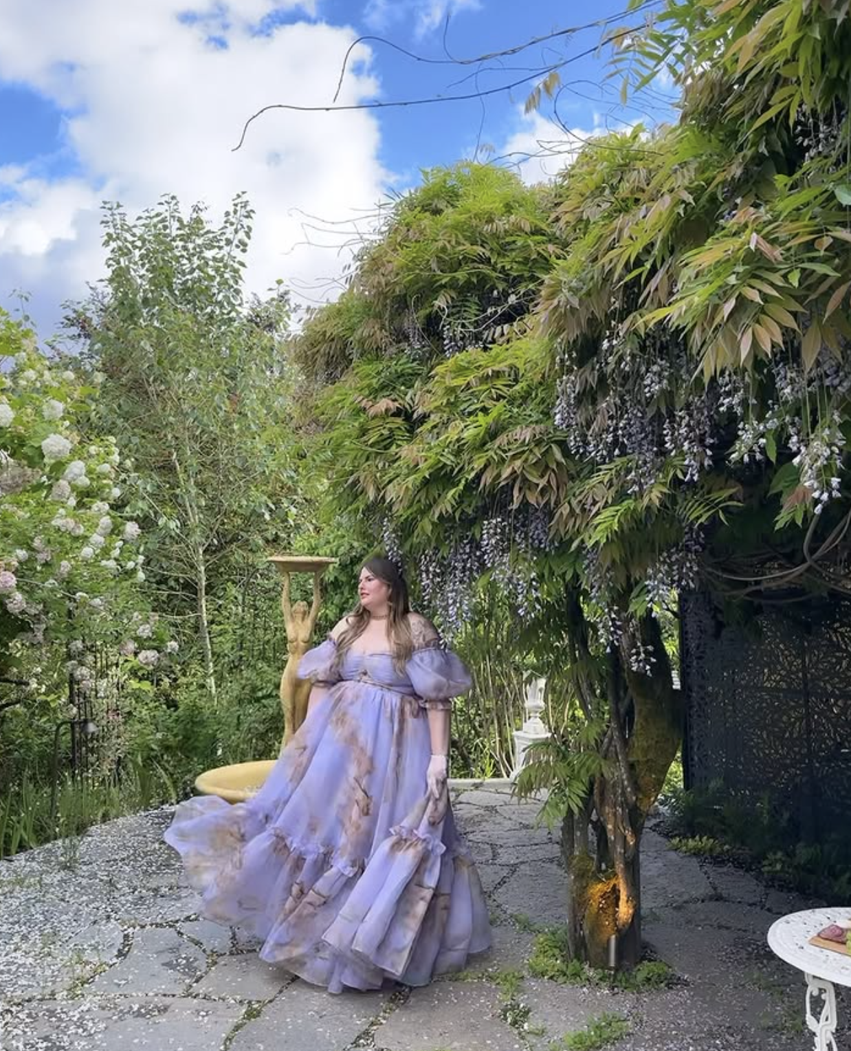 A woman in a flowing lavender dress standing in a lush garden next to a gilded statue and a fountain, with blooming trees and a cloudy blue sky overhead.
