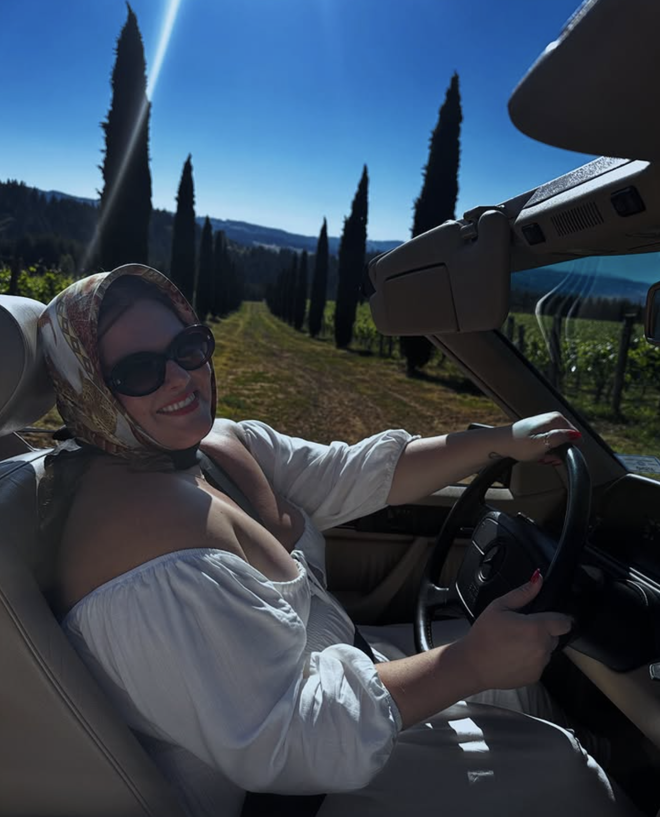 Woman smiling wearing sunglasses and a patterned headscarf, driving a convertible car through a scenic vineyard with tall cypress trees, hills, and clear blue sky in the background.