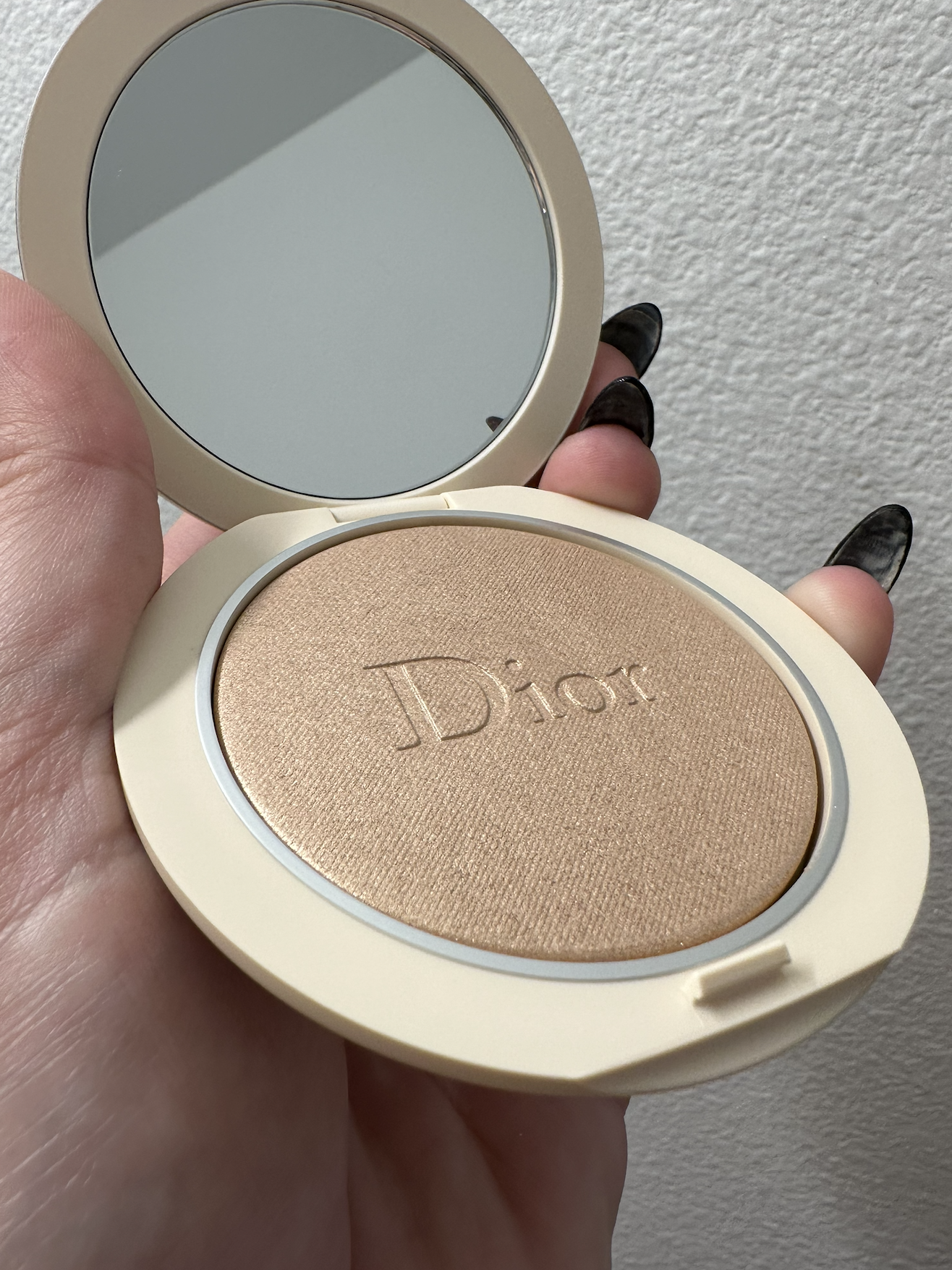 A hand holding an open compact case containing beige-colored Dior pressed powder with the brand name embossed on it, and a mirror reflecting a textured white wall.