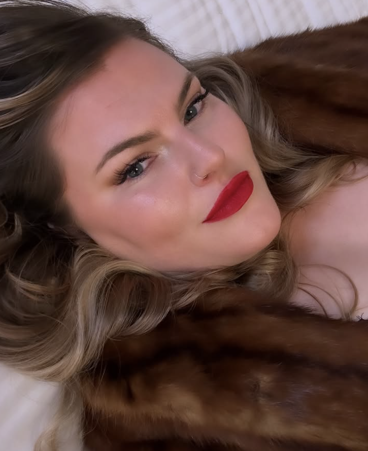 Close-up of a woman with wavy blonde hair, blue eyes, fair skin, red lipstick, and a small nose ring, lying on a bed with brown fur.