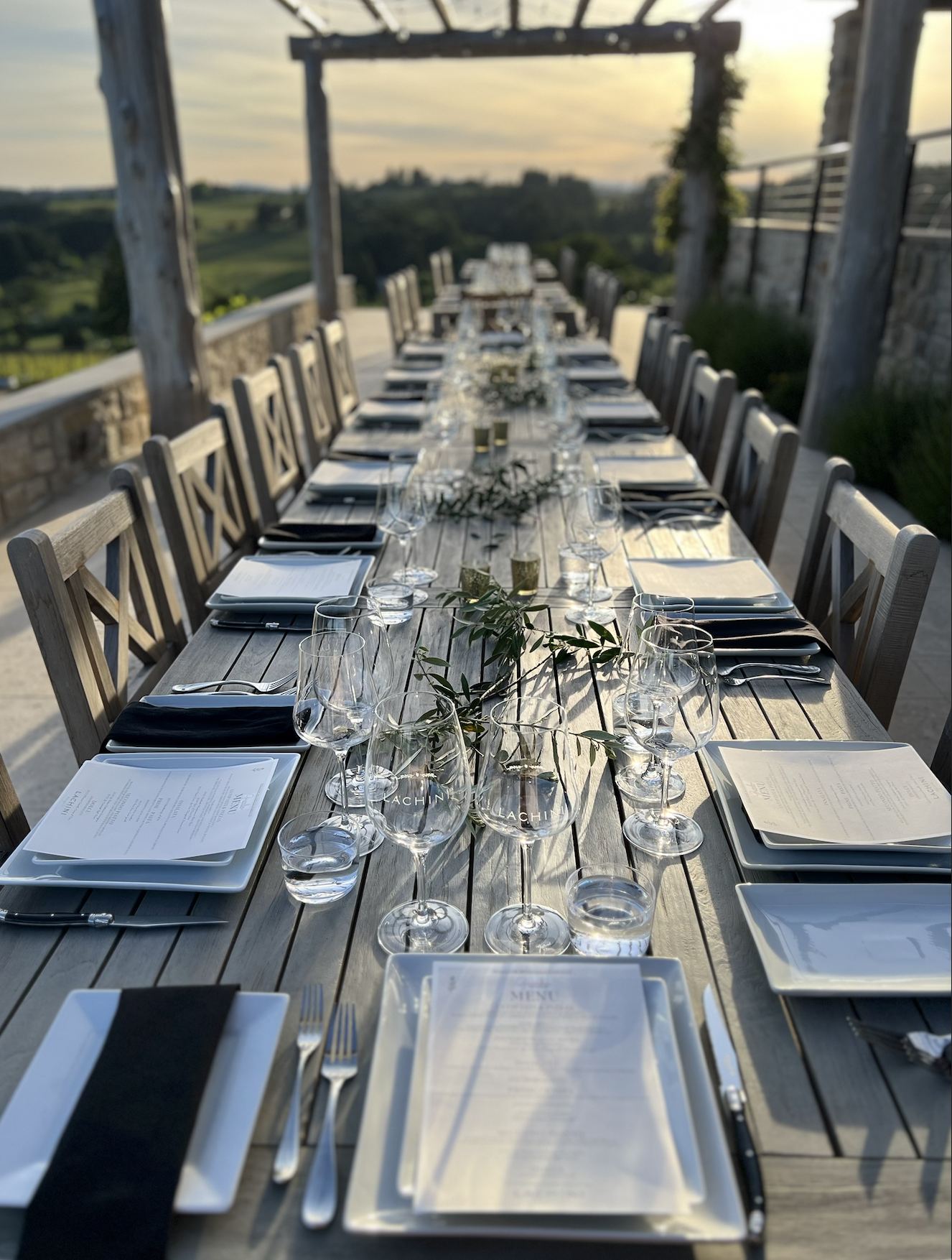 A long outdoor dining table set with wine glasses, water glasses, black napkins, menus, and silverware, situated on a patio with scenic hills in the background during sunset.
