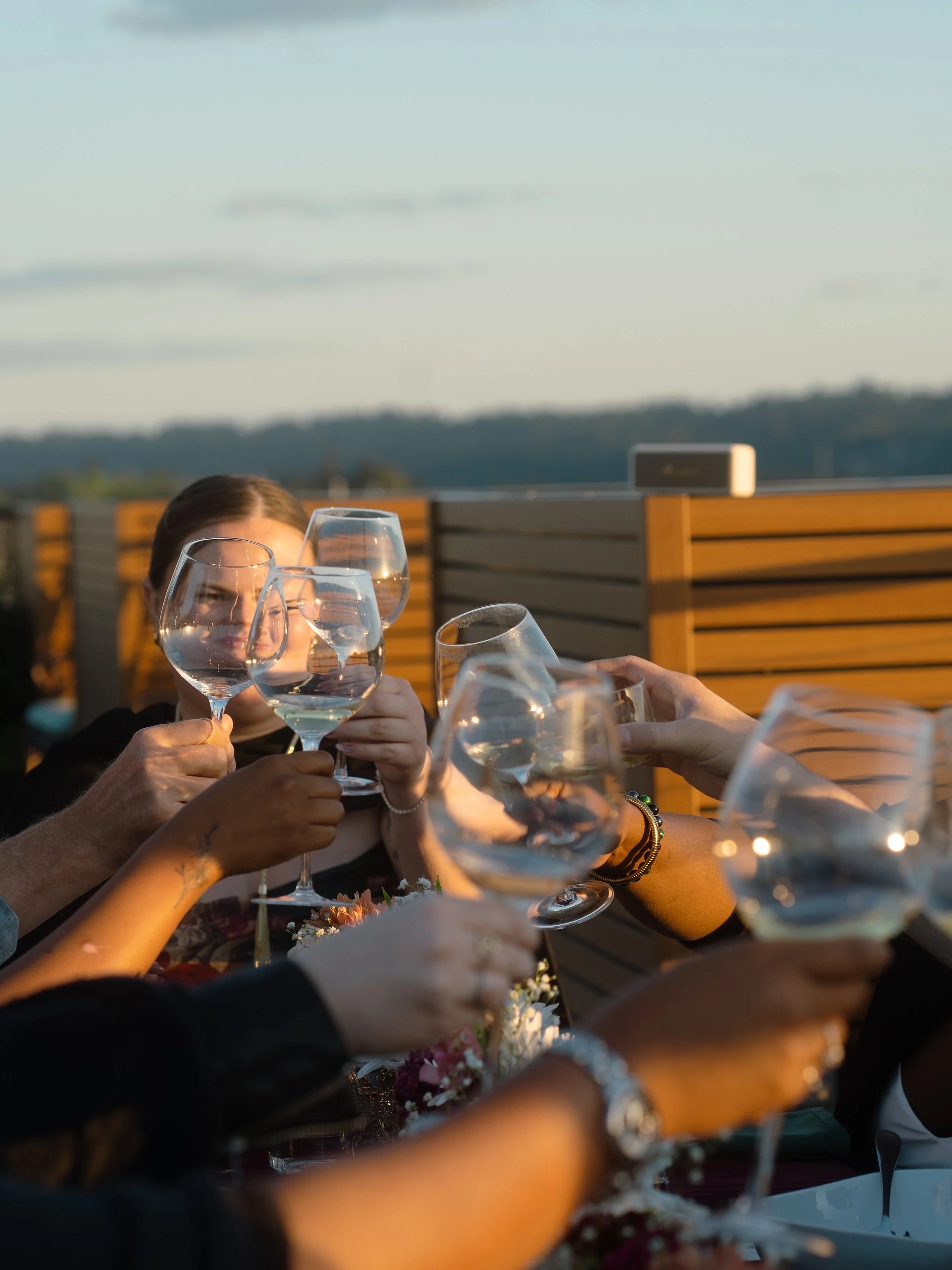 People raising wine glasses in a toast at an outdoor gathering during sunset, with a wooden fence and distant hills in the background.