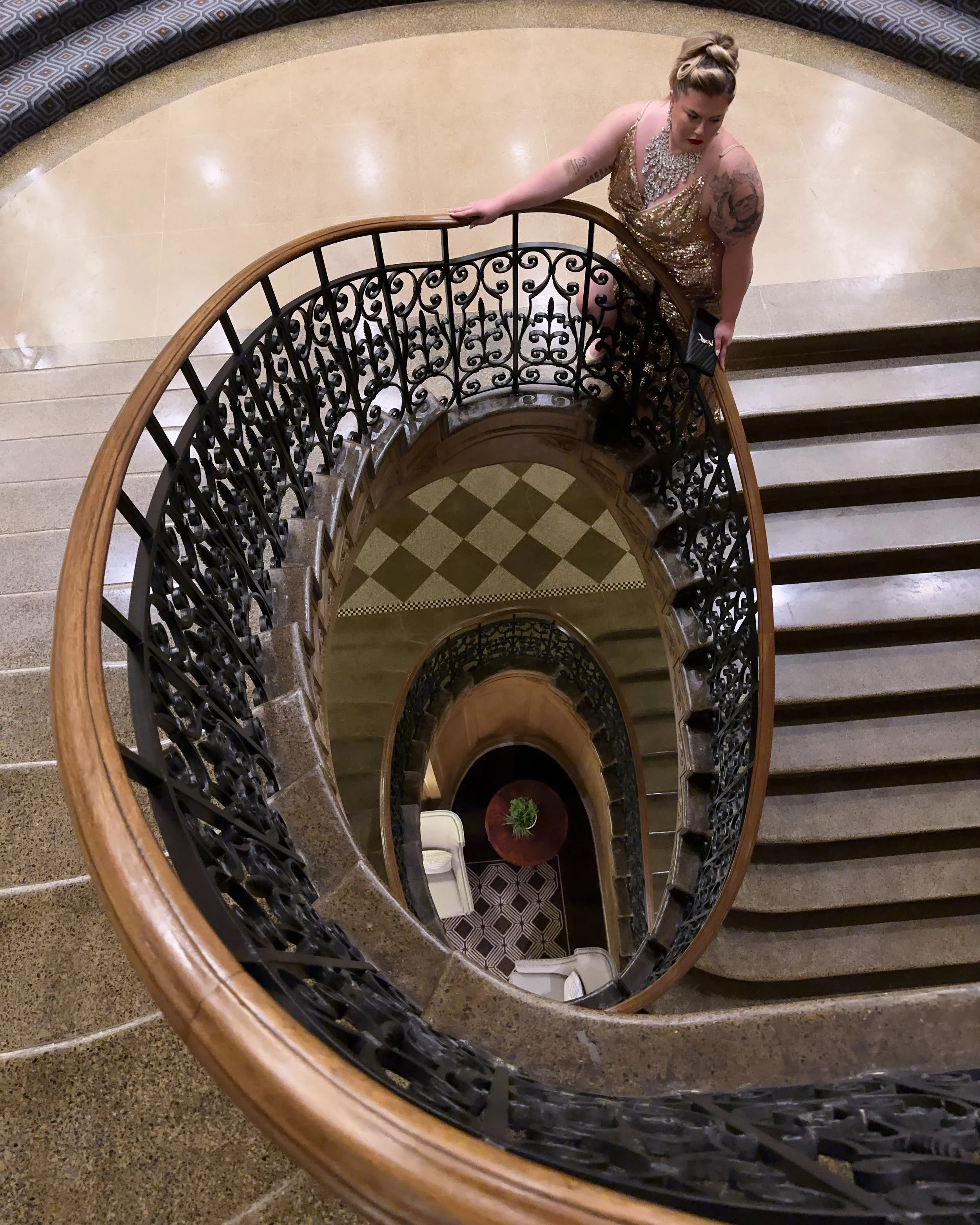 A woman in a gold sequin dress and jewelry, with tattoos, looking down while ascending a spiral staircase with ornate black wrought-iron railing and gray steps, in a building with a beige tiled floor and decorated black and gray patterned wall.