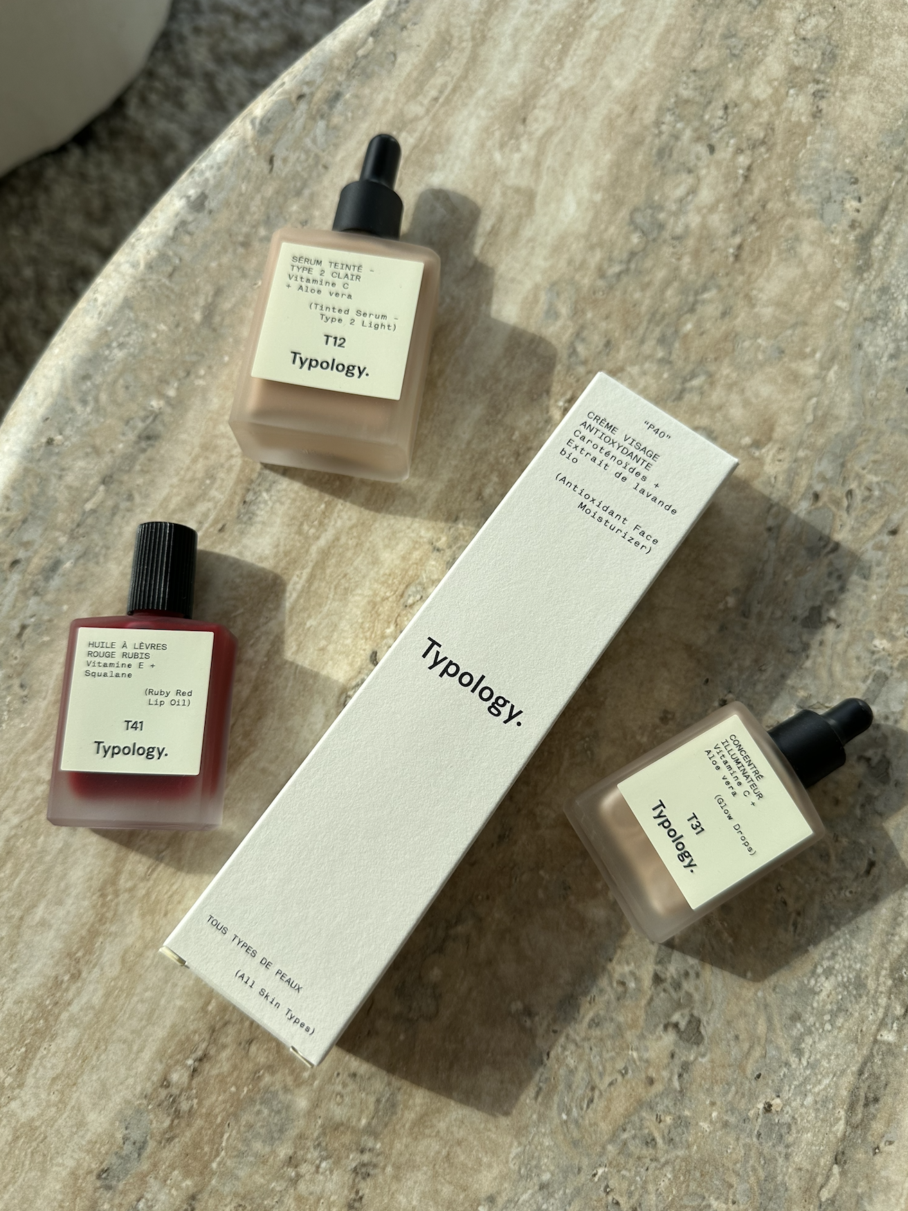 Three skincare serum bottles and a white box on a textured beige surface. The bottles have dropper caps and labels with product details and numbers.