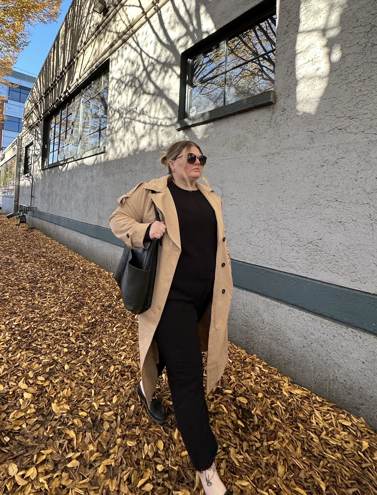 Woman walking on a sidewalk covered with fallen autumn leaves beside a textured gray building with black-trimmed windows, wearing sunglasses, a tan trench coat, black outfit, and carrying a black bag.