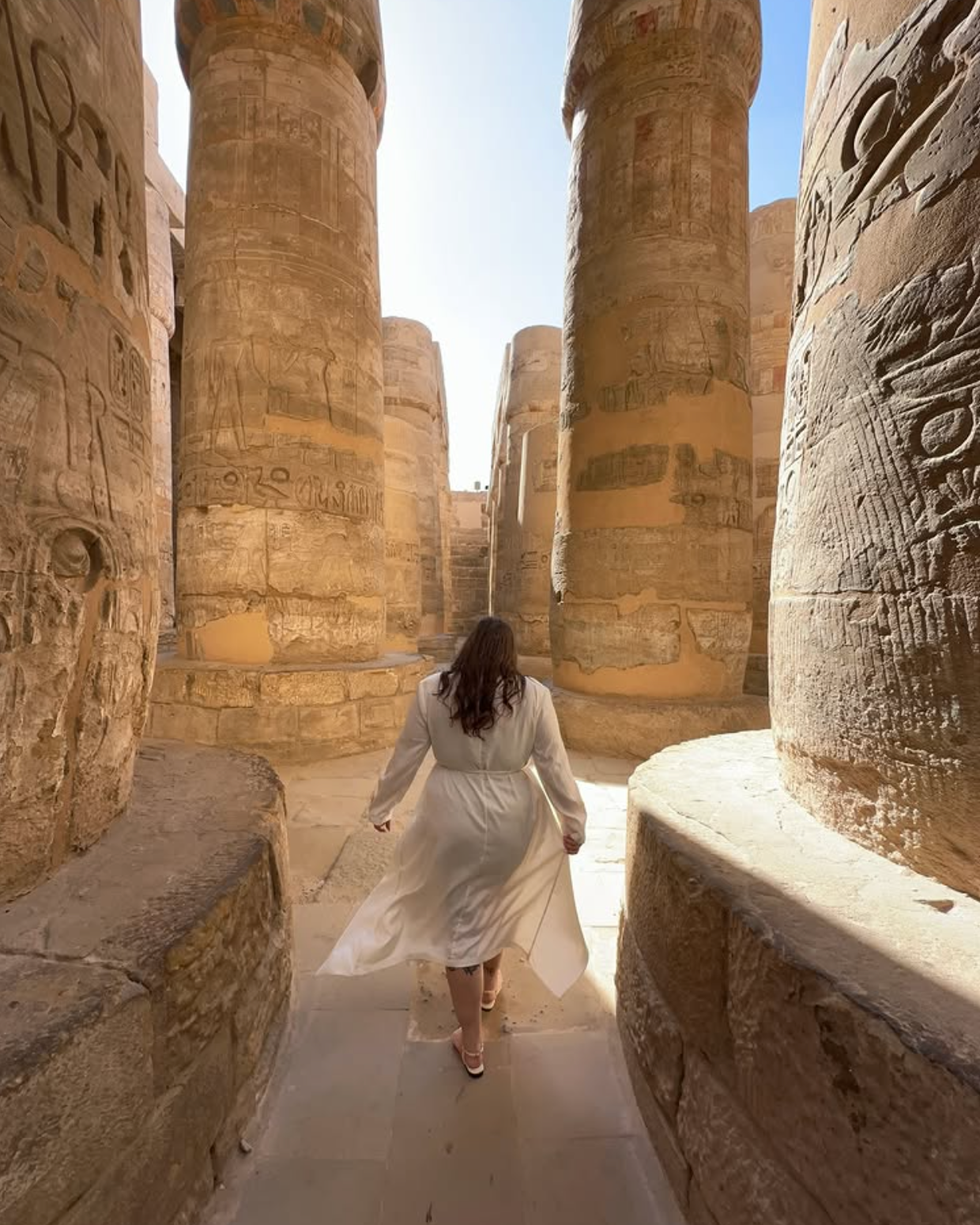 A woman in a white dress walking among ancient Egyptian stone columns with hieroglyphic carvings.