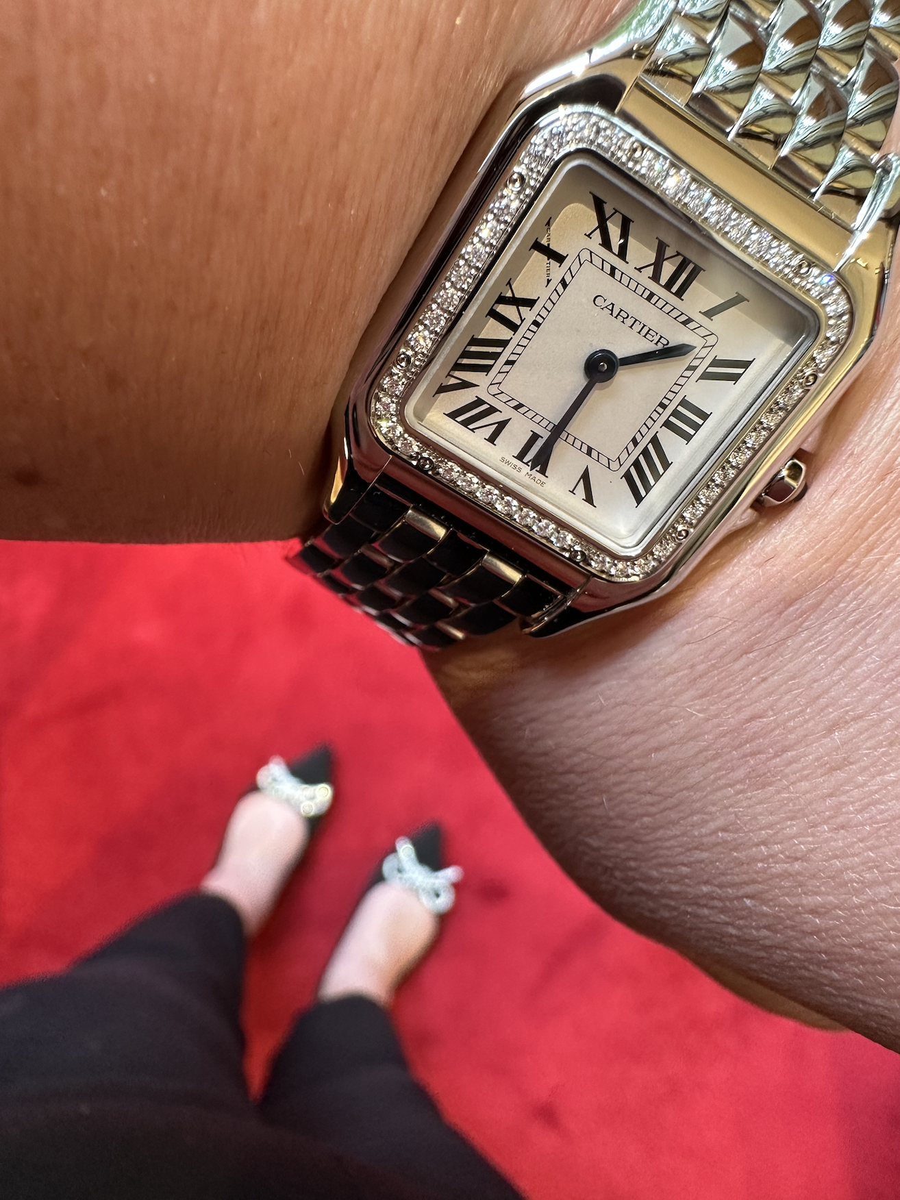 Close-up of a person wearing a Cartier watch with a diamond-encrusted bezel, on their wrist. The person is standing on a red carpet, with their feet visible wearing black shoes with glittery bows.