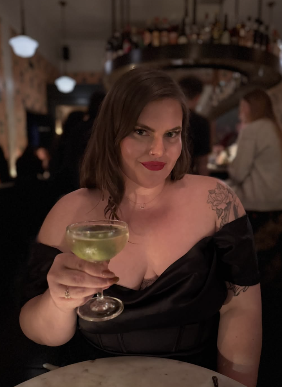 A woman with shoulder-length brown hair and red lipstick, holding a cocktail glass filled with a light-colored drink, smiling at a restaurant or bar with warmly lit ambiance.