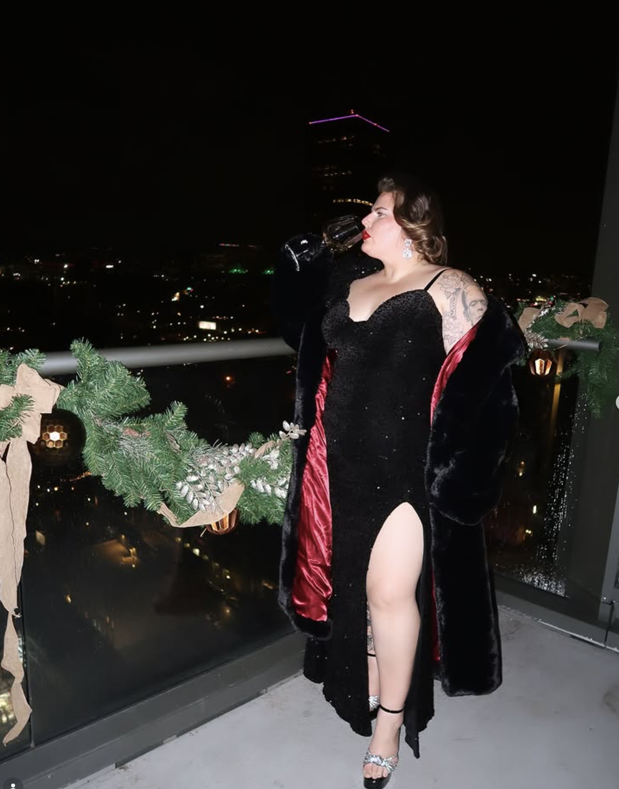 A woman dressed in black with a red lining, wearing a black dress with a high slit, stands on a balcony at night, drinking from a wine glass, with city lights and a tall building with pink lights in the background, decorated with green Christmas garland and ornaments.