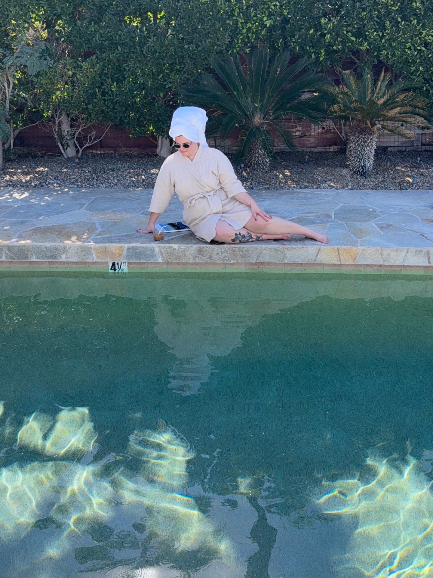 Person sitting on the poolside, wearing a towel on their head and sunglasses, next to a swimming pool with clear water, greenery in the background, and a height marker reading 4½ feet.