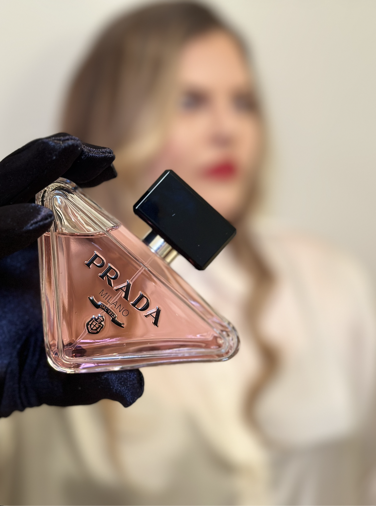 A woman with blurred features holding a Prada perfume bottle with a black cap and pink liquid, wearing black gloves, against a light background.