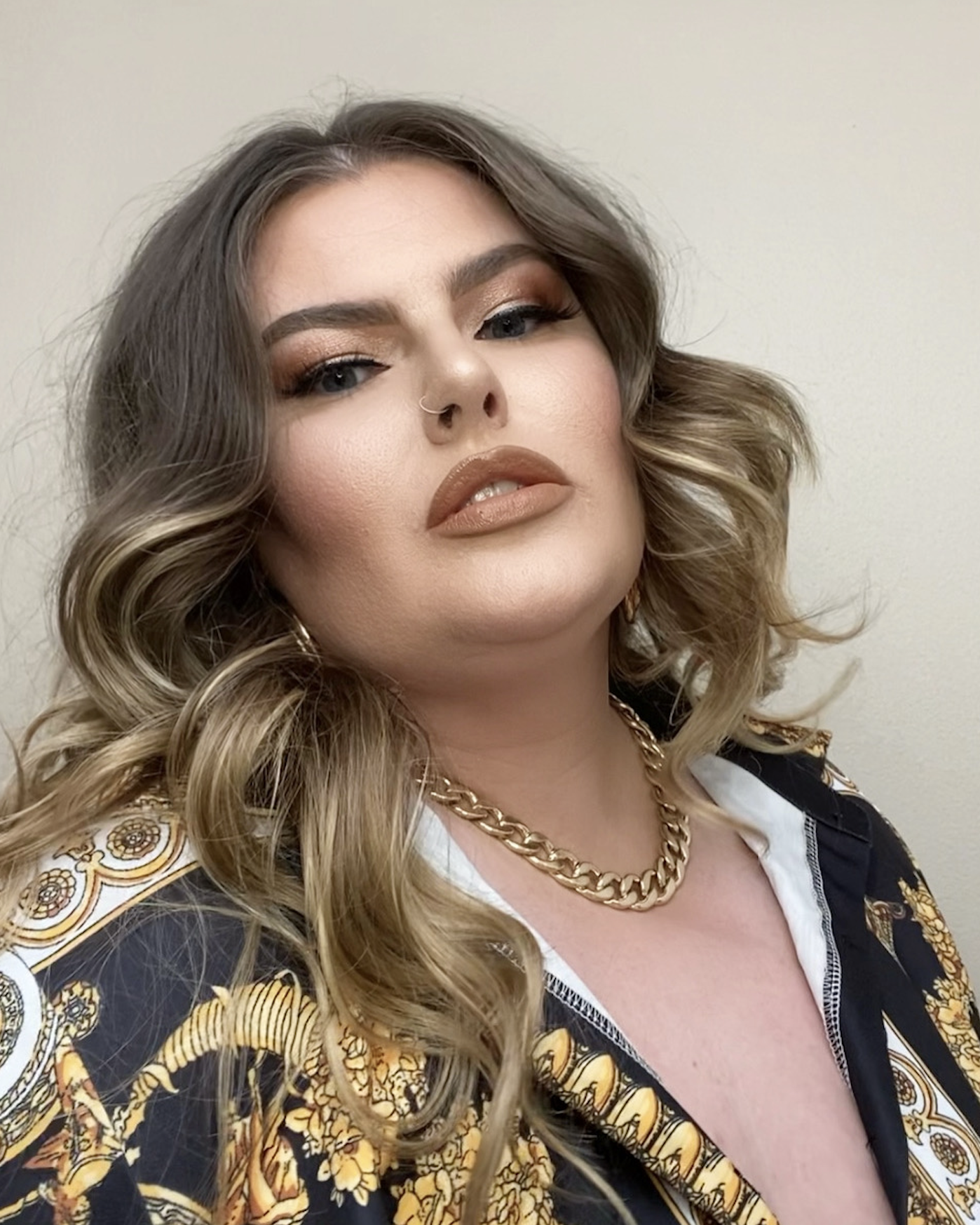 A woman with wavy blonde hair, makeup, and a nose ring, wearing a gold chain necklace and a patterned black and gold top, taking a selfie against a plain background.