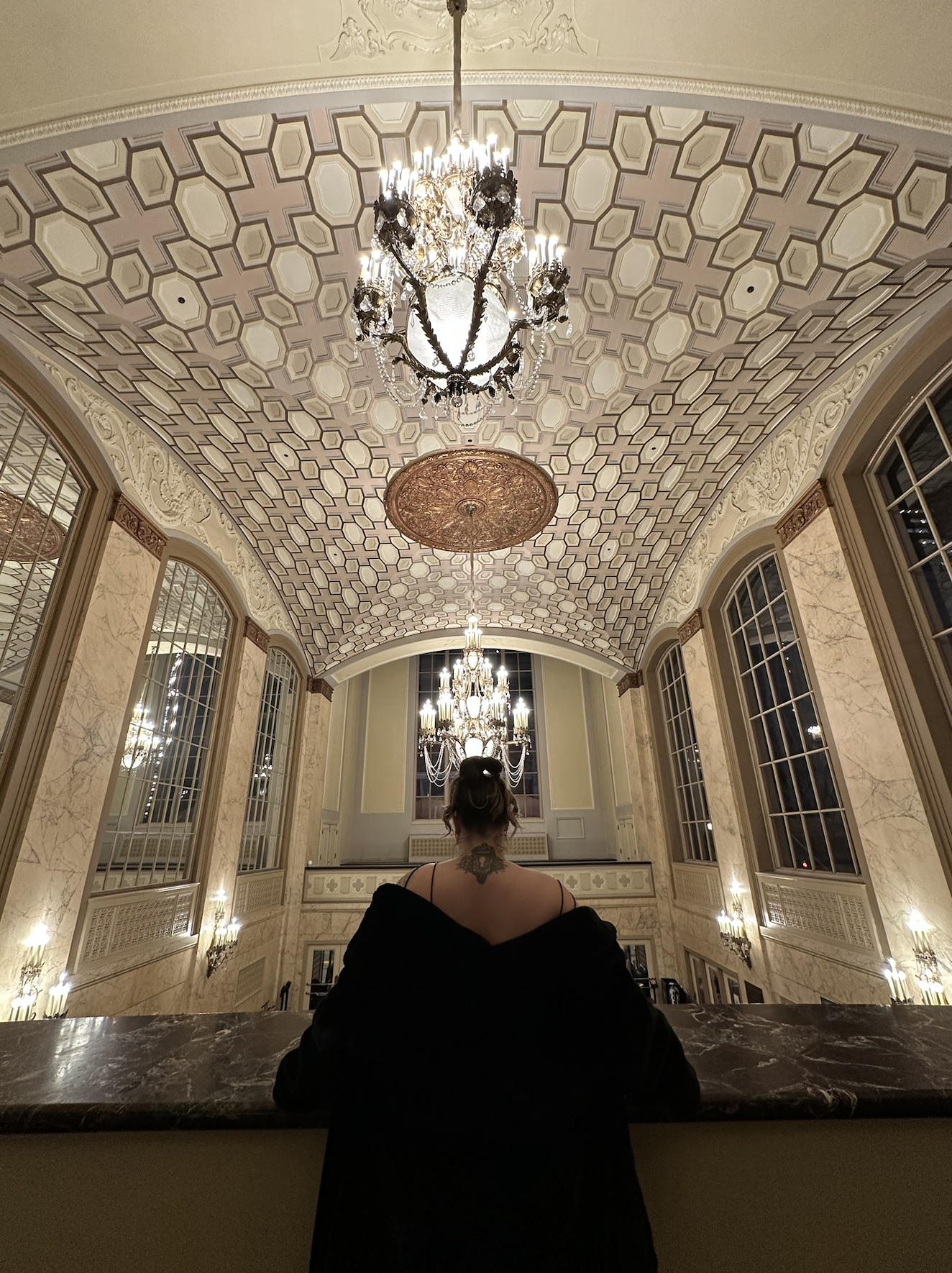 Interior of an elegant hall with a high, decorated ceiling and ornate chandeliers, large arched windows, marble walls, and a person with dark hair and tattoos sitting at a marble counter, facing away from the camera.