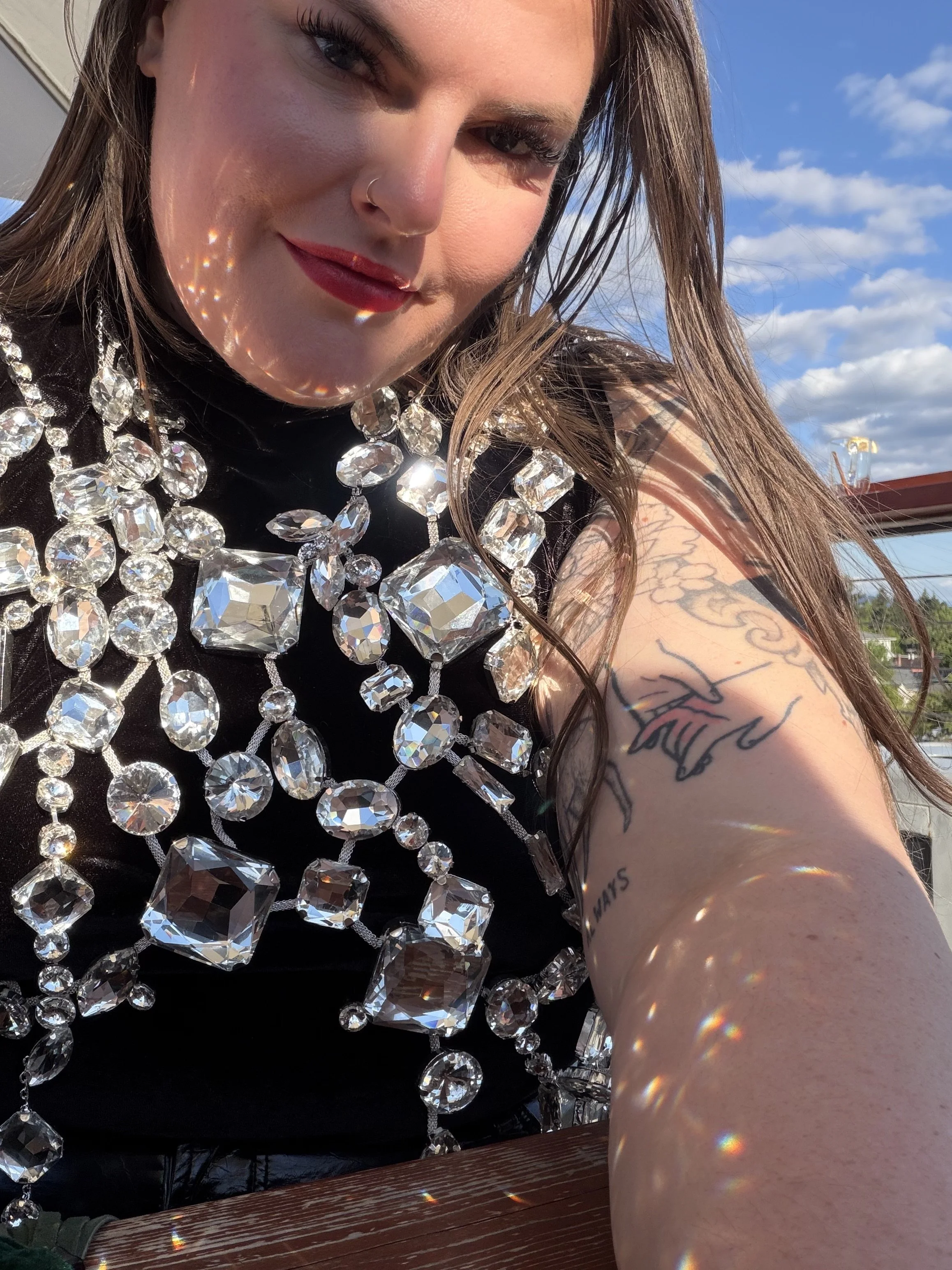 A woman with long brown hair, wearing a large, sparkling rhinestone necklace, taking a selfie outdoors against a blue sky with scattered clouds. She has light makeup with red lipstick, a nose piercing, and tattoos on her right arm.