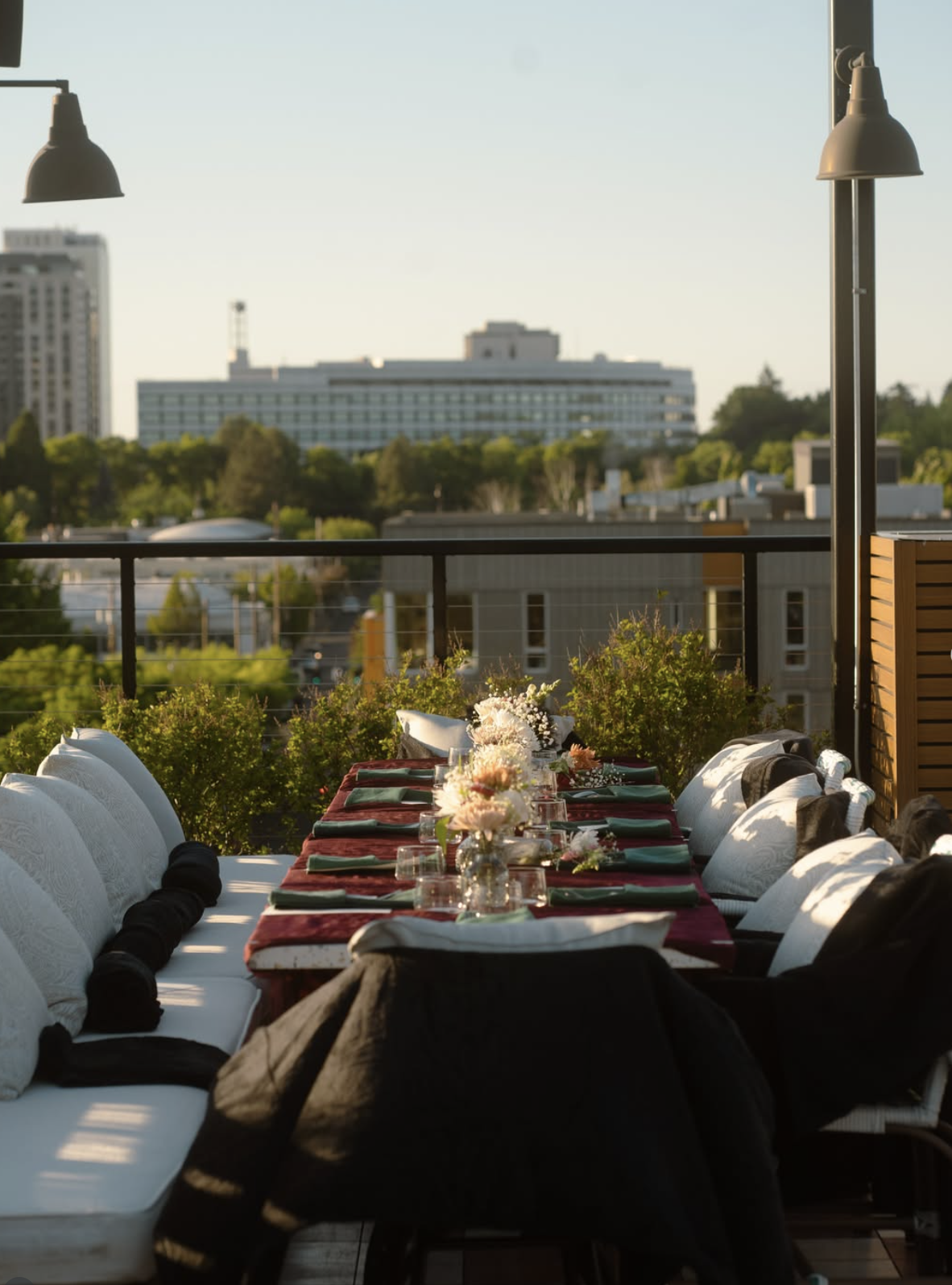 A rooftop dinner setup with a long table decorated with flowers and place settings, overlooking a cityscape with buildings and greenery in the background during daylight.