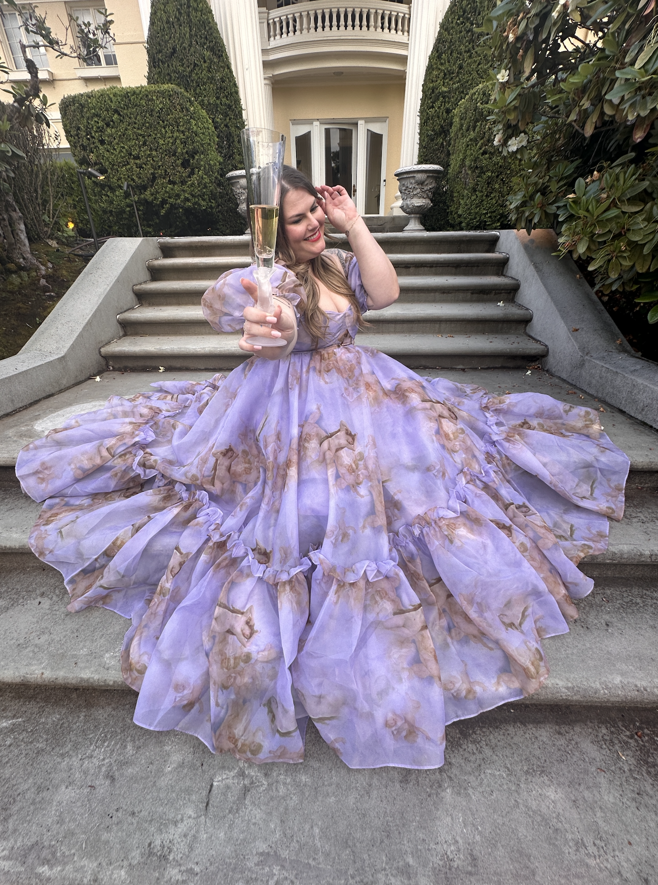 A woman sitting on outdoor steps in a flowing, pastel dress with a horse print, holding a champagne flute and smiling