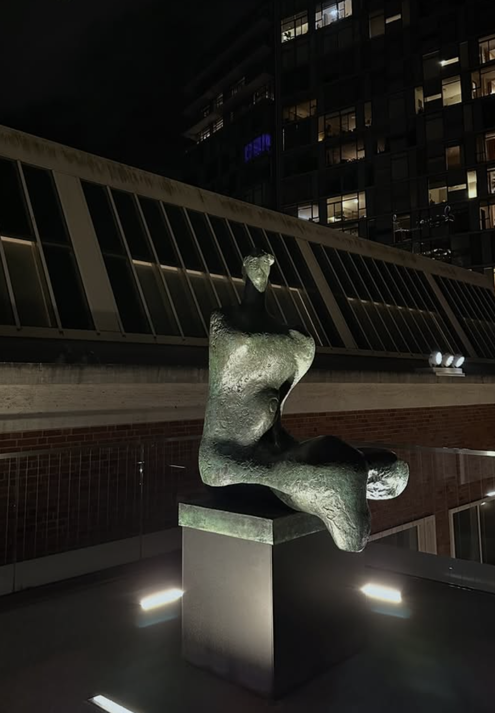Nighttime view of a modern apartment building with illuminated windows, featuring an abstract sculpture of a seated human figure on a pedestal outside, illuminated by ground lighting.