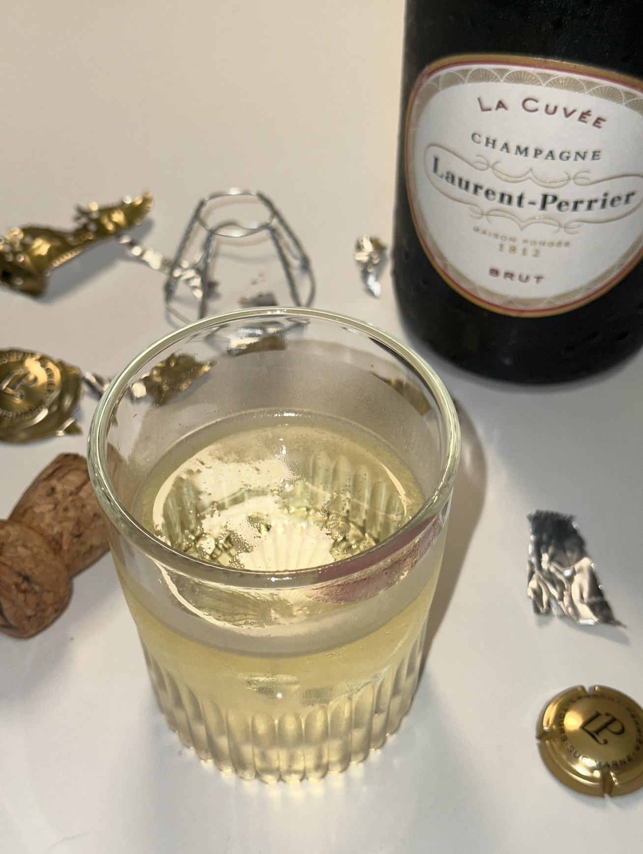 A glass of champagne with a bottle of Laurent-Perrier champagne in the background, alongside corks, foil wrappers, and gold and silver decorative items on a white surface.