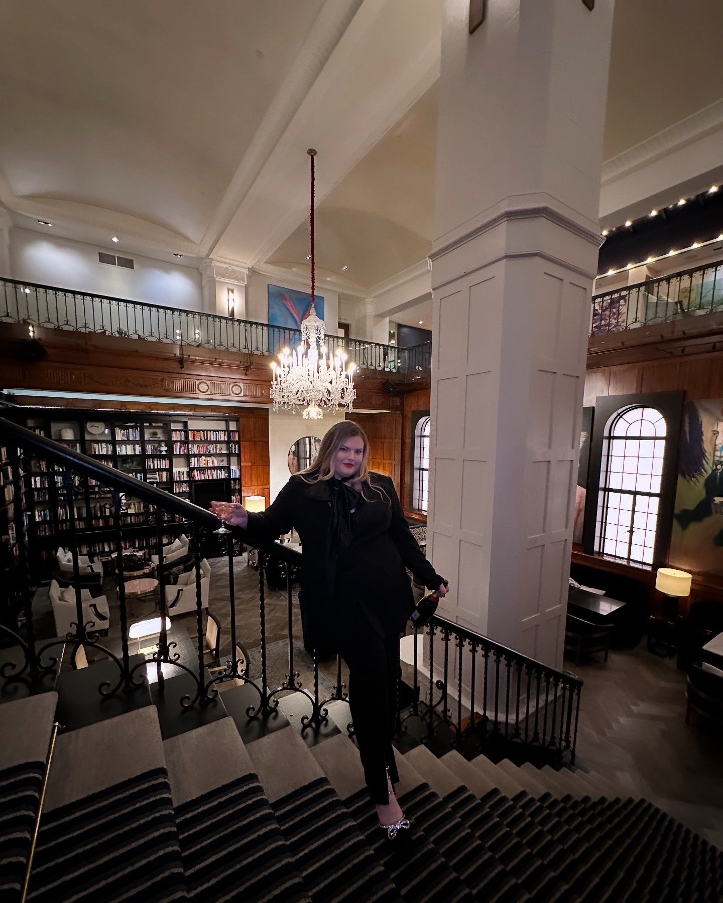 A woman with blonde hair in a black outfit standing on a staircase in an elegant, well-lit room with a chandelier, bookshelves, and decorative art on the walls.