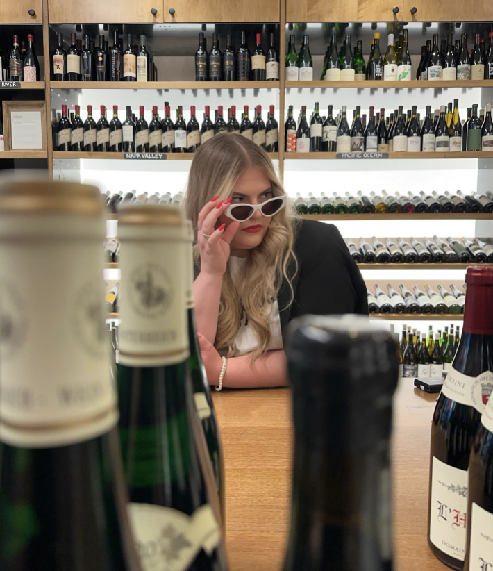 A young woman with long blonde hair and red lipstick is sitting at a wooden table surrounded by bottles of wine, looking through her white sunglasses in a wine shop or cellar.