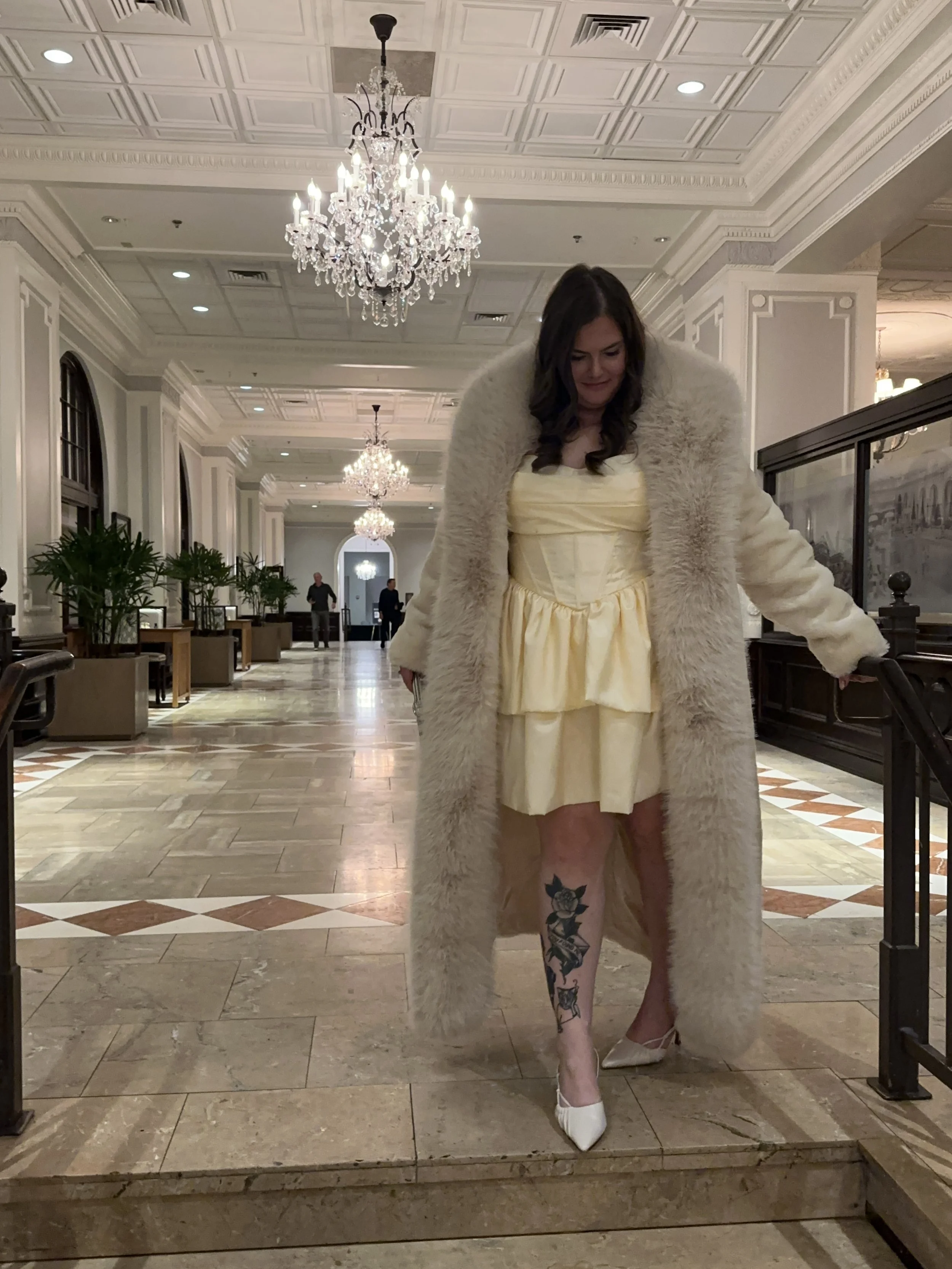 A woman wearing a yellow dress and a long white fur coat, standing on a marbled staircase in a luxurious and elegant hall with high ceilings, chandeliers, and decorative molding.